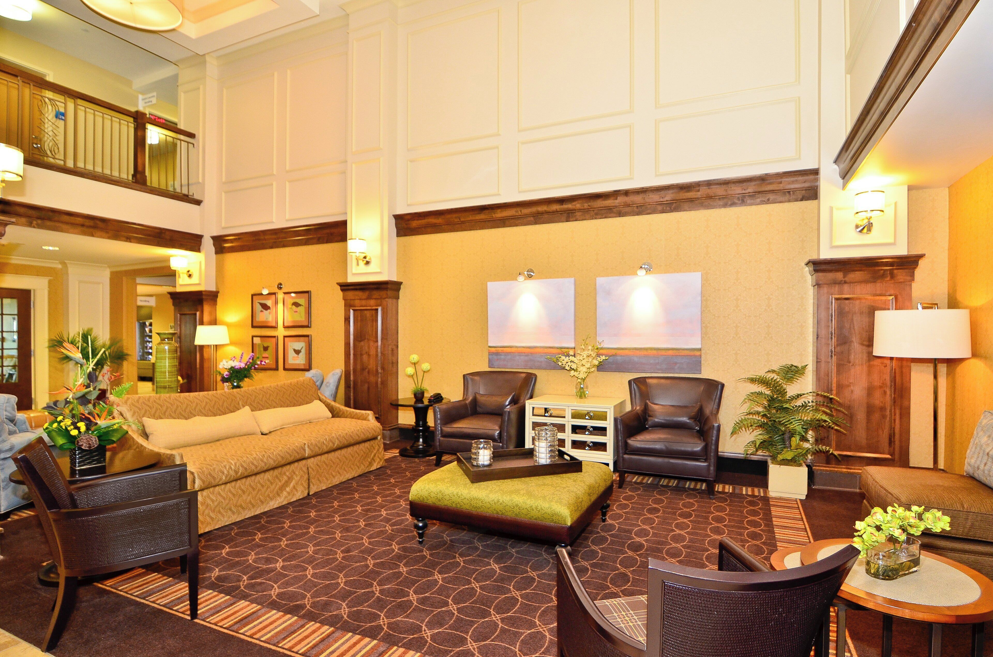 A spacious lobby with a high ceiling, a central seating area with a sofa and chairs, a coffee table, and a decorative vase with flowers.