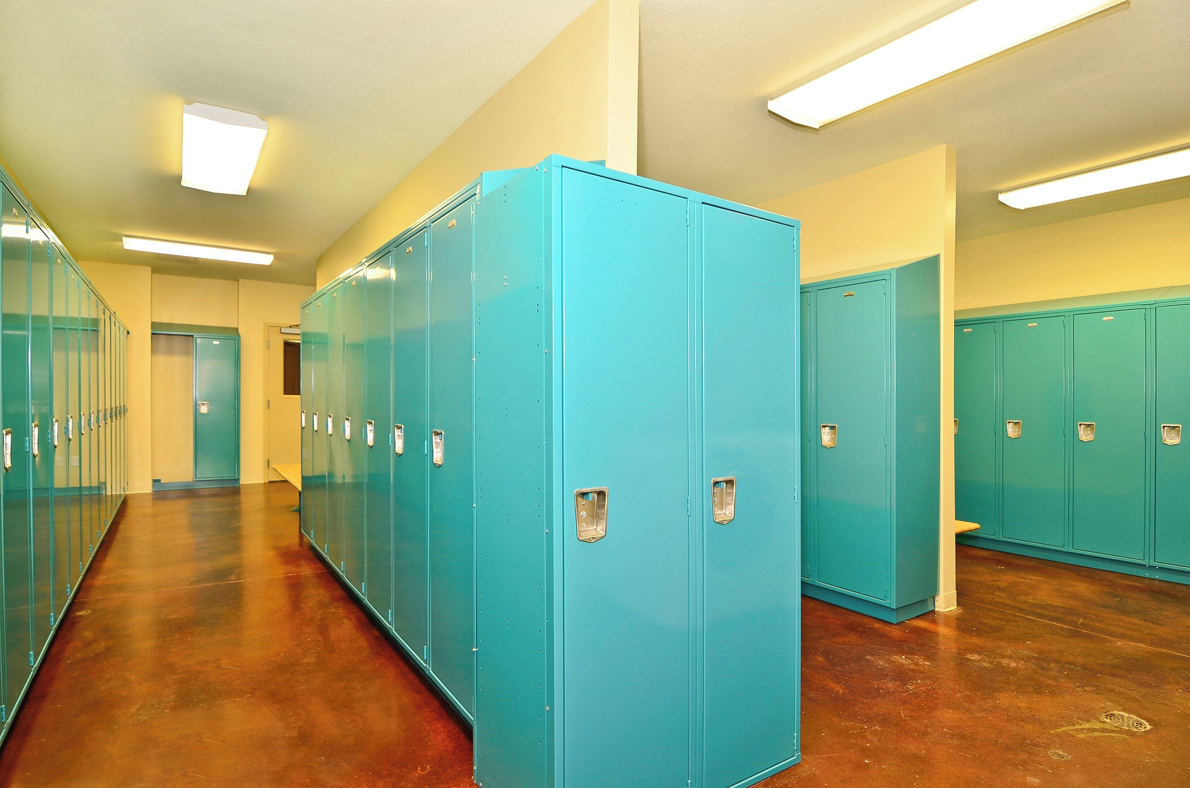 A hallway with rows of teal lockers, wooden flooring, and fluorescent ceiling lights.
