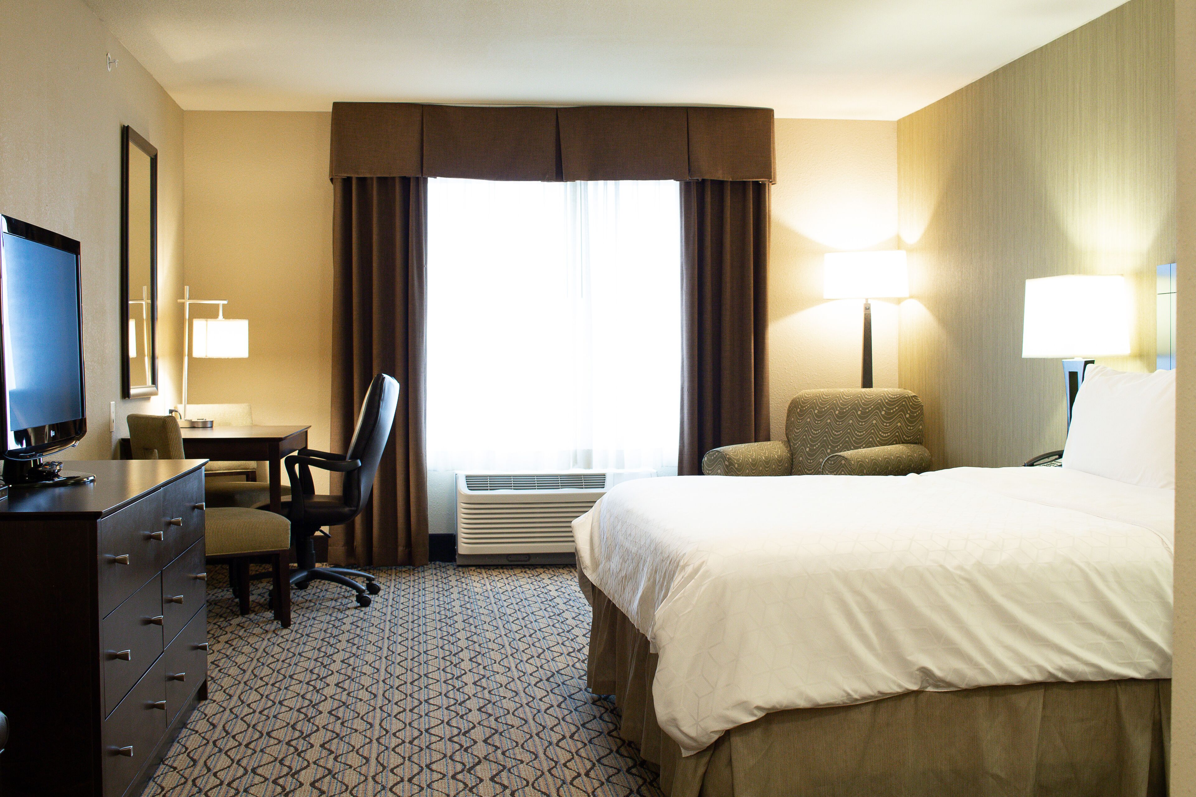 A hotel room with a bed, a desk with a computer, a chair, a dresser, and a window with curtains.