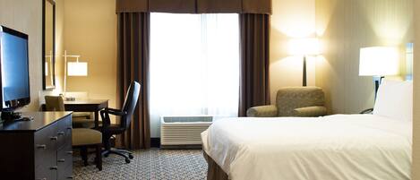 A hotel room with a bed, a desk with a computer, a chair, a dresser, and a window with curtains.