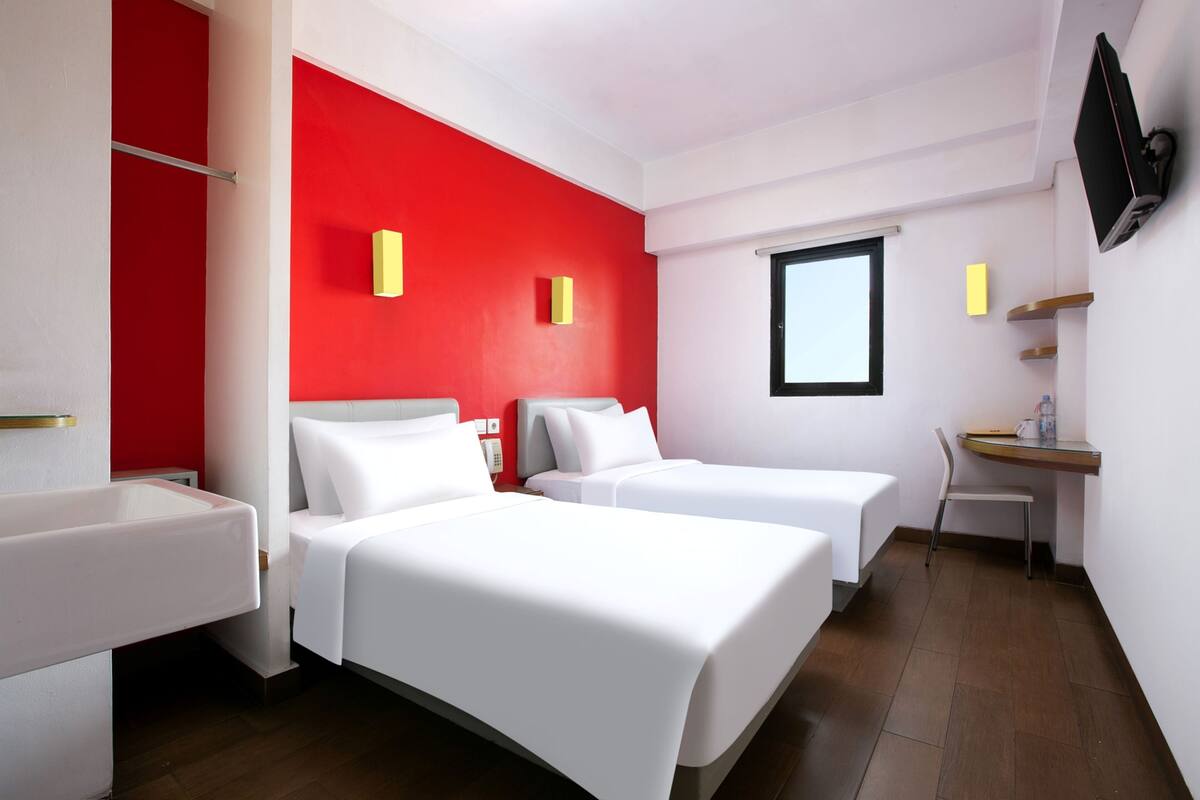 A hotel room with two beds, a desk, a chair, a TV mounted on the wall, and a window.