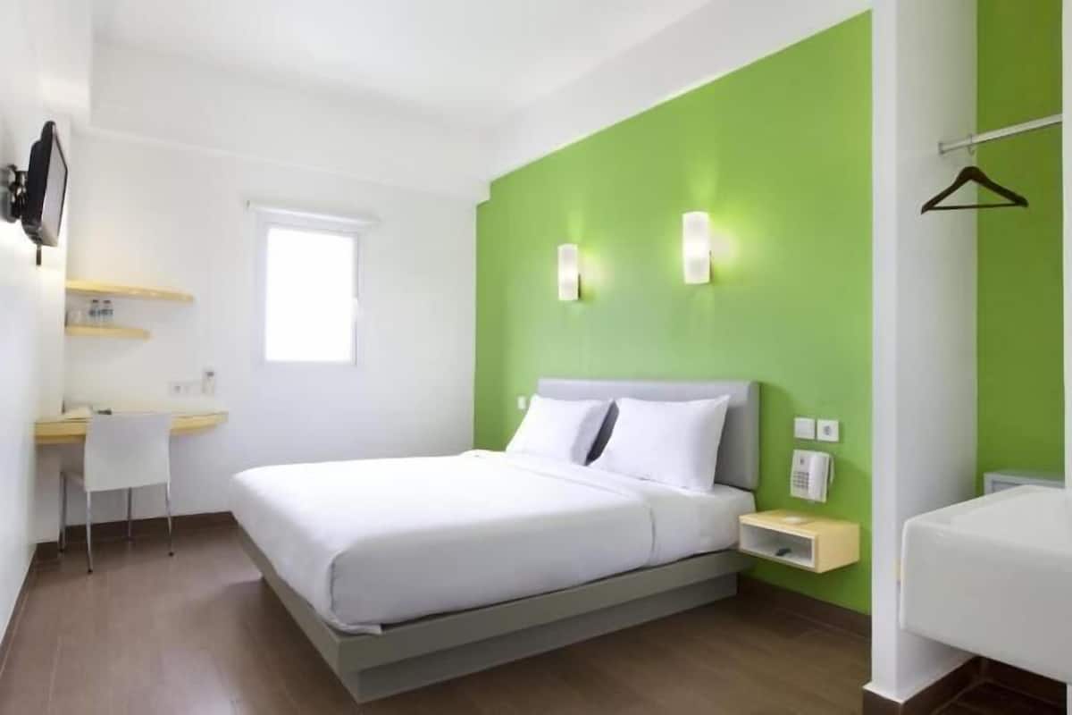 A modern hotel room with a bed, bedside table, a small desk with a chair, a window, and a green accent wall.