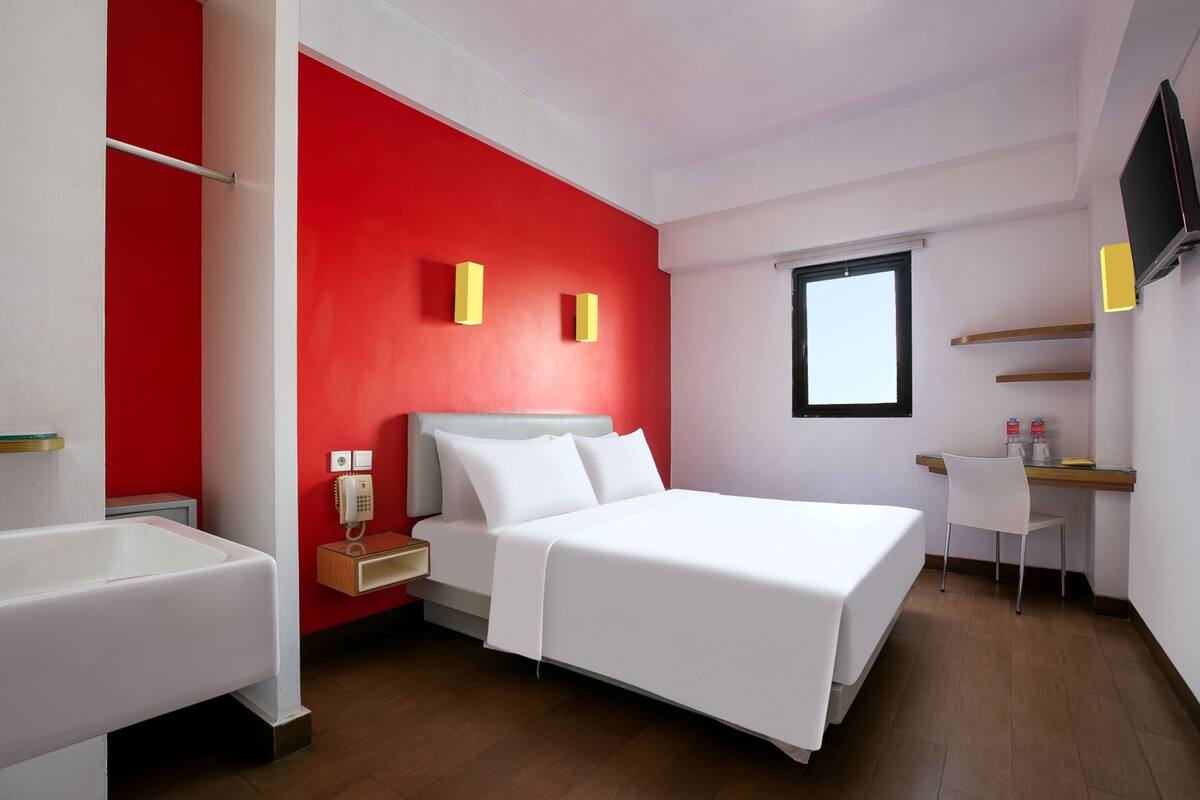 A hotel room with a bed, a desk, a chair, a window, and a red accent wall.