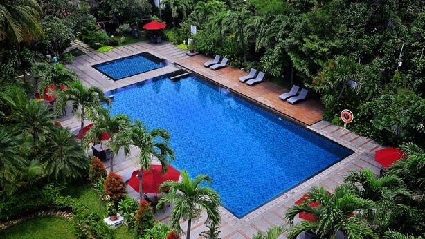 Una gran piscina rectangular con cubierta de madera y sillones de descanso, rodeada de exuberante vegetación y palmeras.