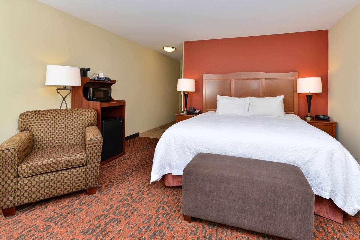 A hotel room with a bed, a chair, a desk with a microwave, and a nightstand with lamps.