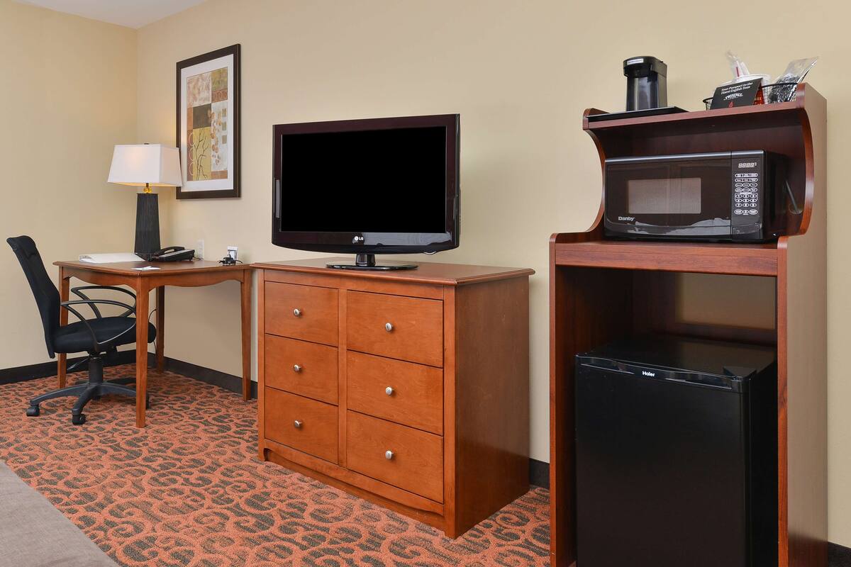 A hotel room with a wooden desk, a flat-screen TV, a microwave, a refrigerator, a chair, and a lamp.