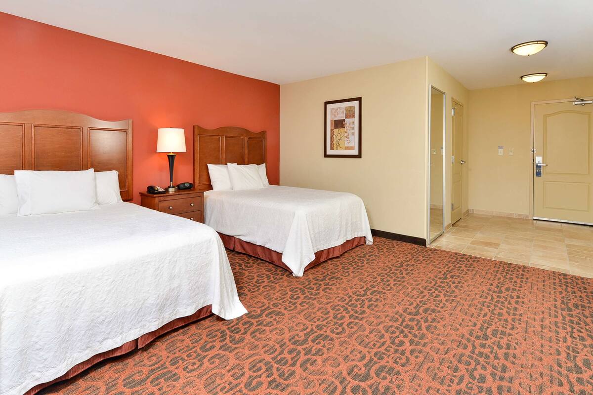 A hotel room with two beds, a nightstand with a lamp, a framed picture on the wall, and a door leading to a bathroom.