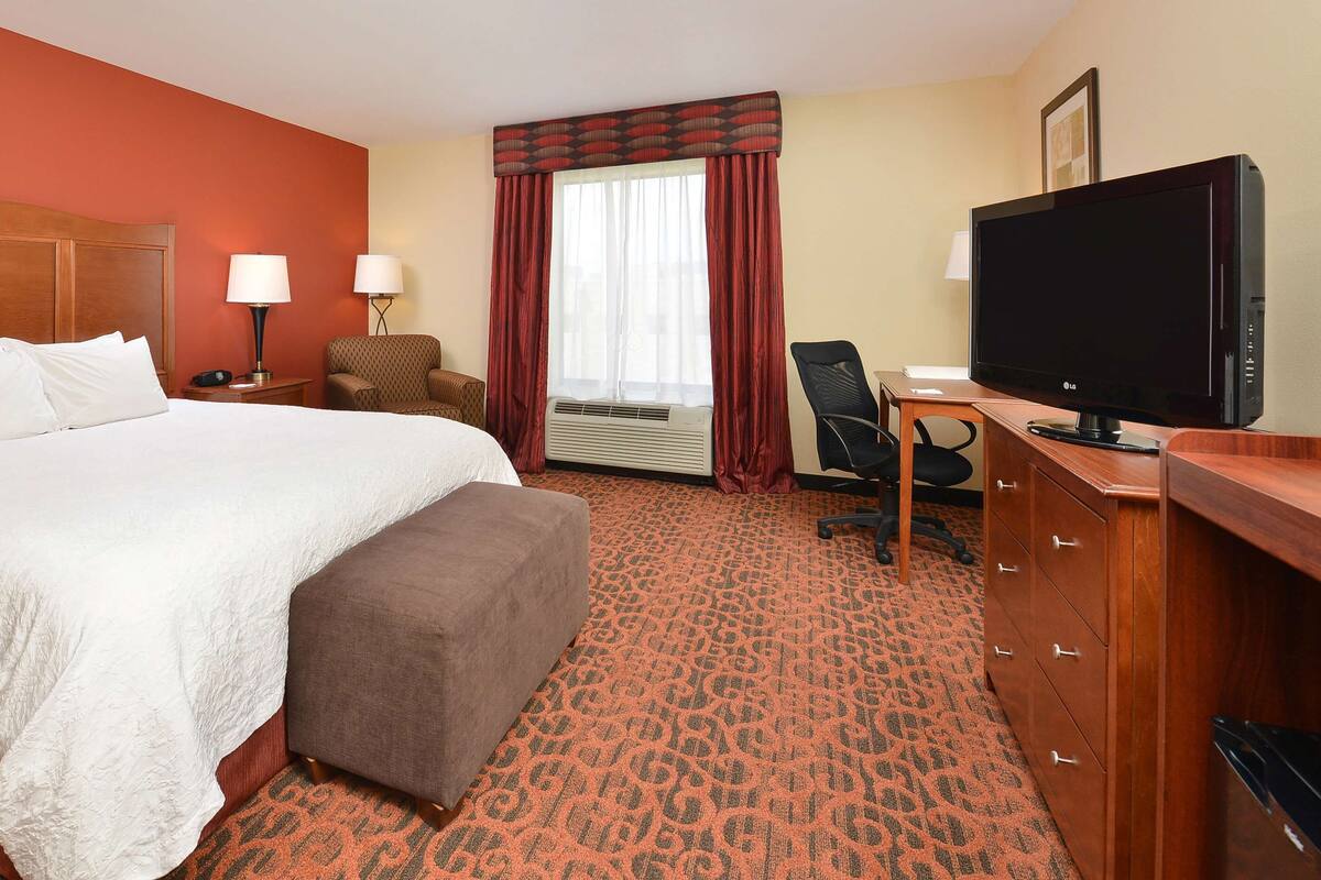A hotel room with a large bed, a desk with a flat-screen TV, a chair, a desk lamp, a telephone, and a wall-mounted air conditioner.