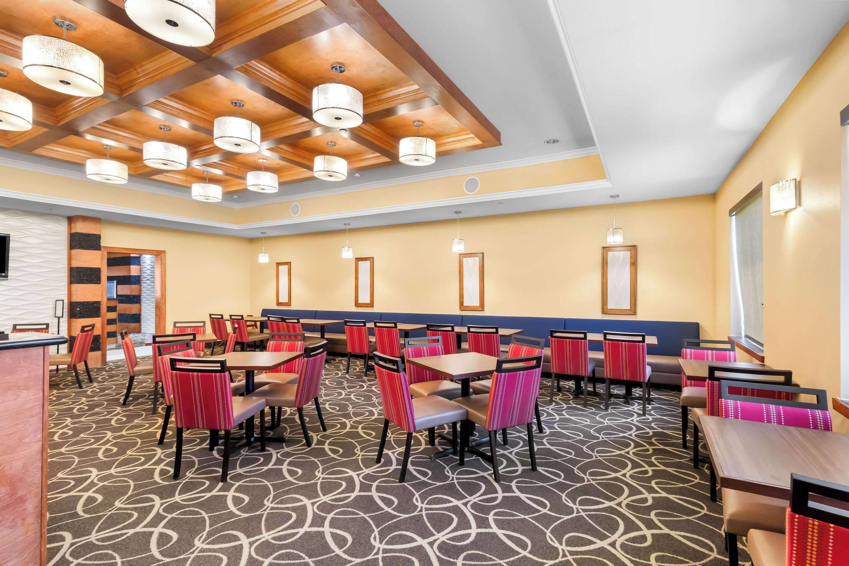 A hotel dining area with multiple tables and chairs, a patterned carpet, and ceiling lights.