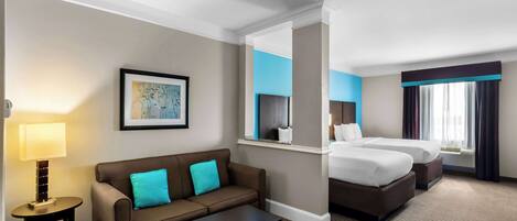 A hotel room with a sofa, two beds, a small table, a lamp, and a framed picture on the wall.