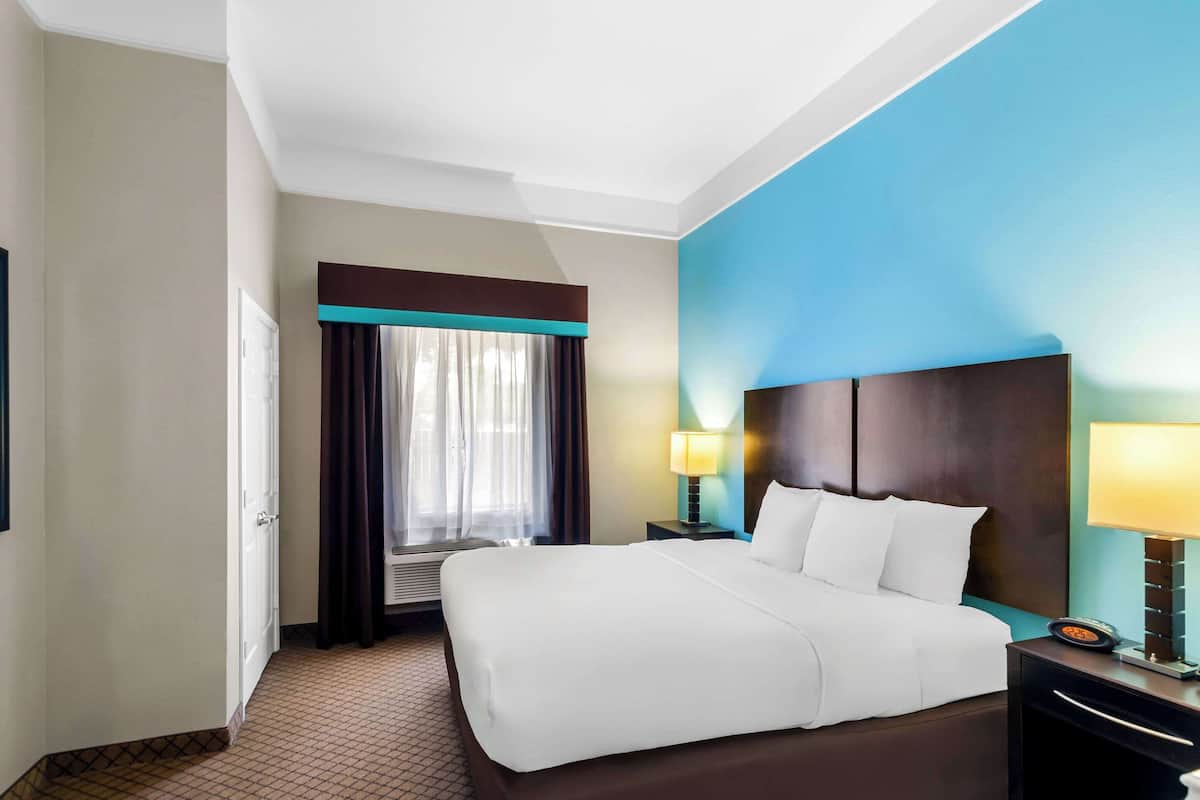 A hotel room with a large bed, two bedside tables with lamps, a window with curtains, and a door.
