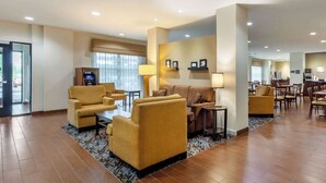 Eine geräumige Lobby mit einer Couch, Sesseln und einem Couchtisch. Es gibt eine Rezeption, einen Essbereich und ein großes Fenster mit Jalousien.