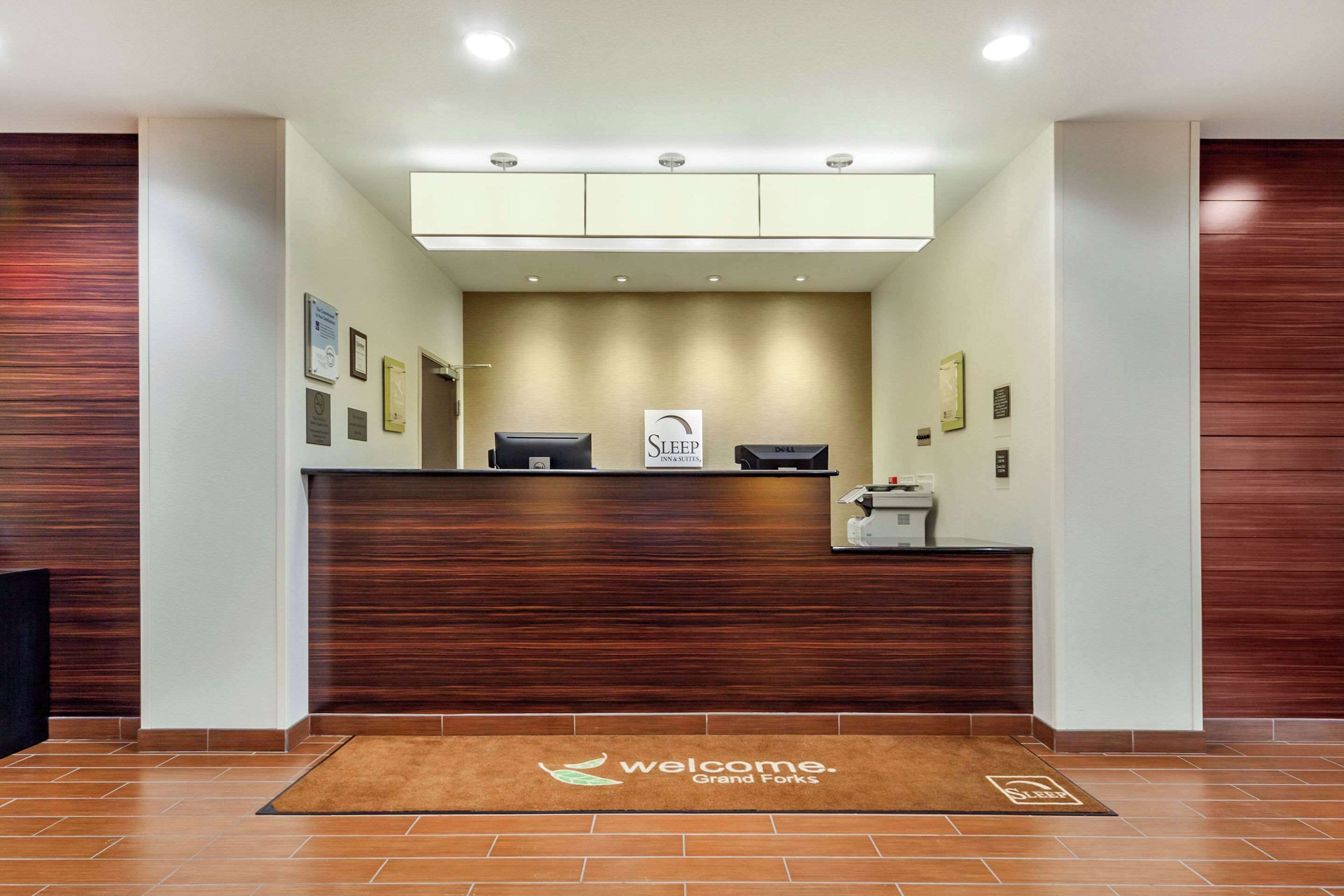 A reception area with a wooden reception desk, a 'Welcome' mat, and framed certificates on the wall.