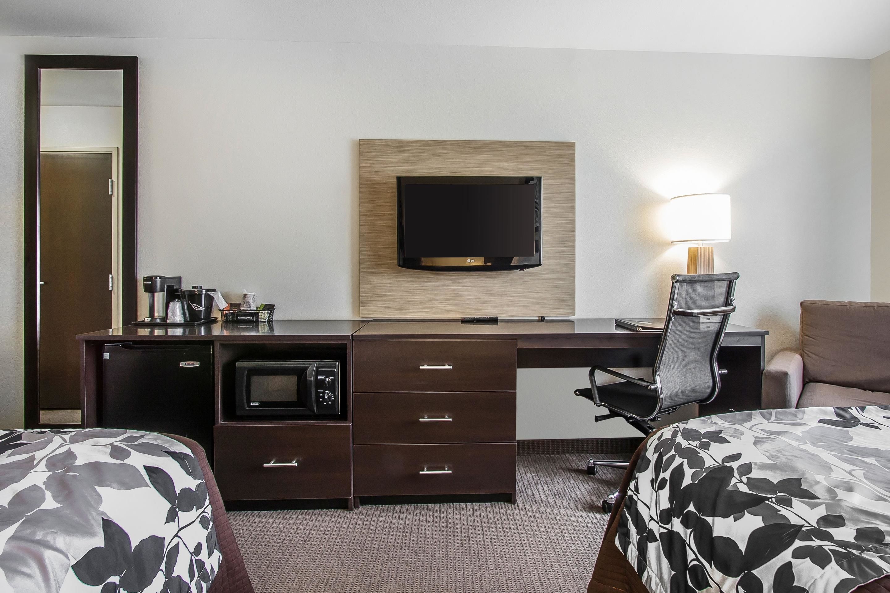 Habitación de hotel con escritorio, televisor de pantalla plana en la pared, microondas, cafetera, lámpara y dos camas con ropa de cama estampada.
