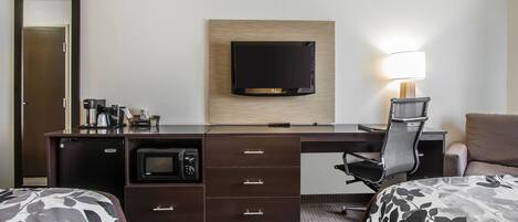 A hotel room with a desk, a flat-screen TV mounted on the wall, a microwave, a coffee maker, a lamp, and two beds with floral bedding.