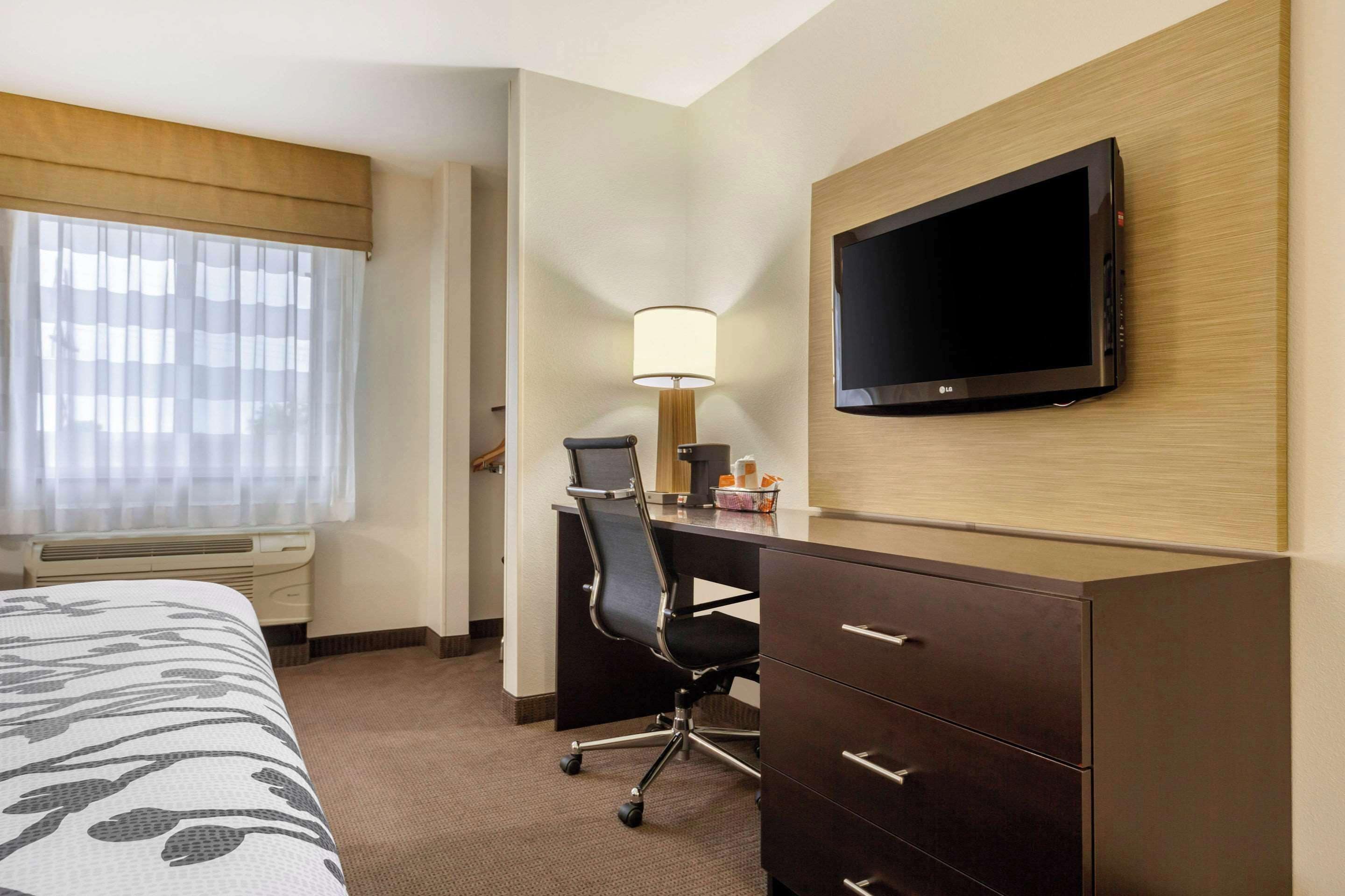 A hotel room with a bed, a desk with a chair, a TV mounted on the wall, and a lamp.