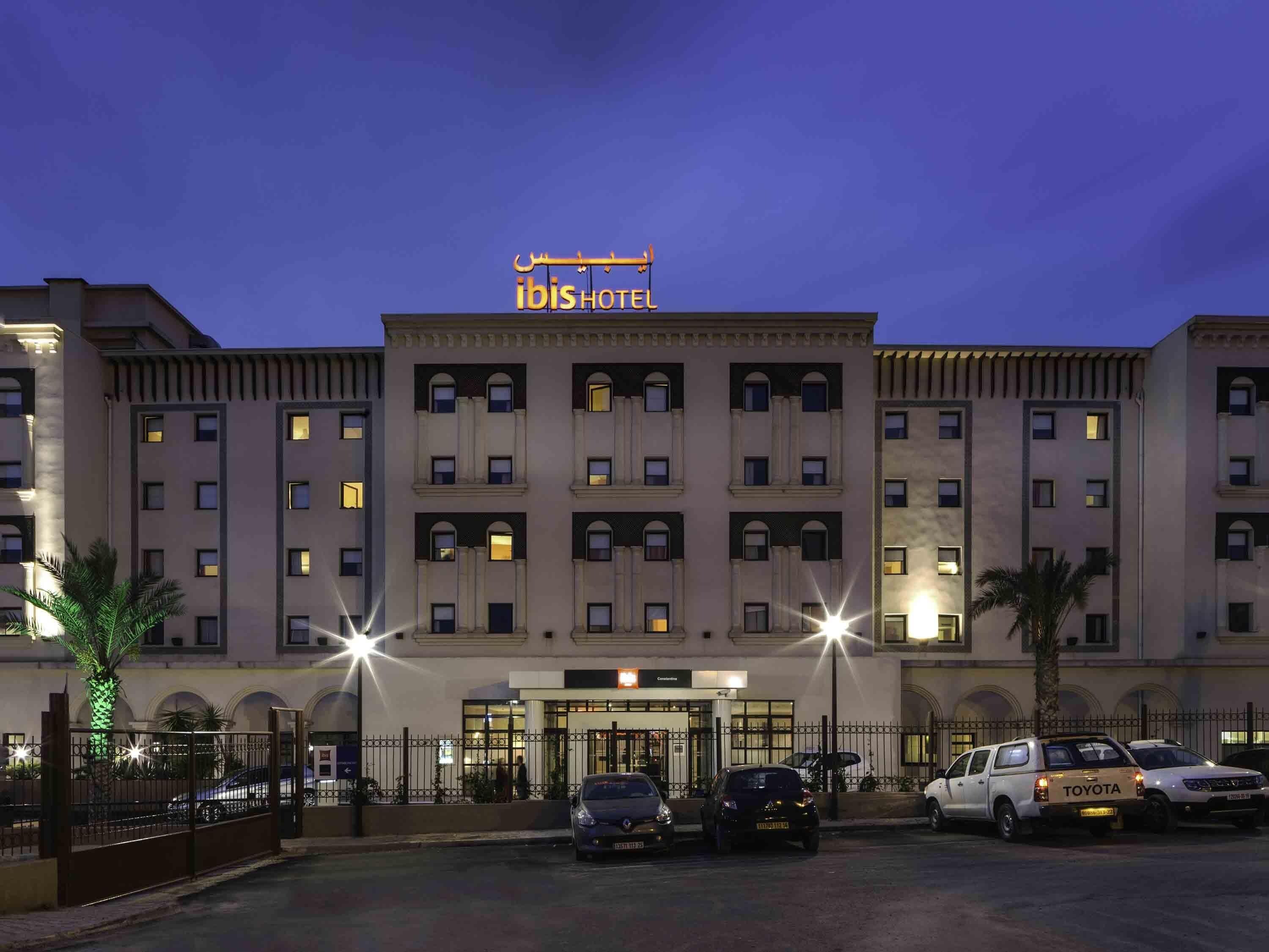 Et hotel med en markant indgang, flere etager og en parkeringsplads med biler.