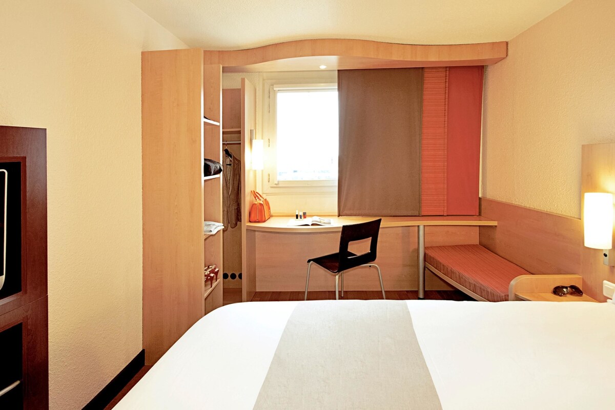 A compact hotel room with a bed, a desk, a chair, and a wardrobe.