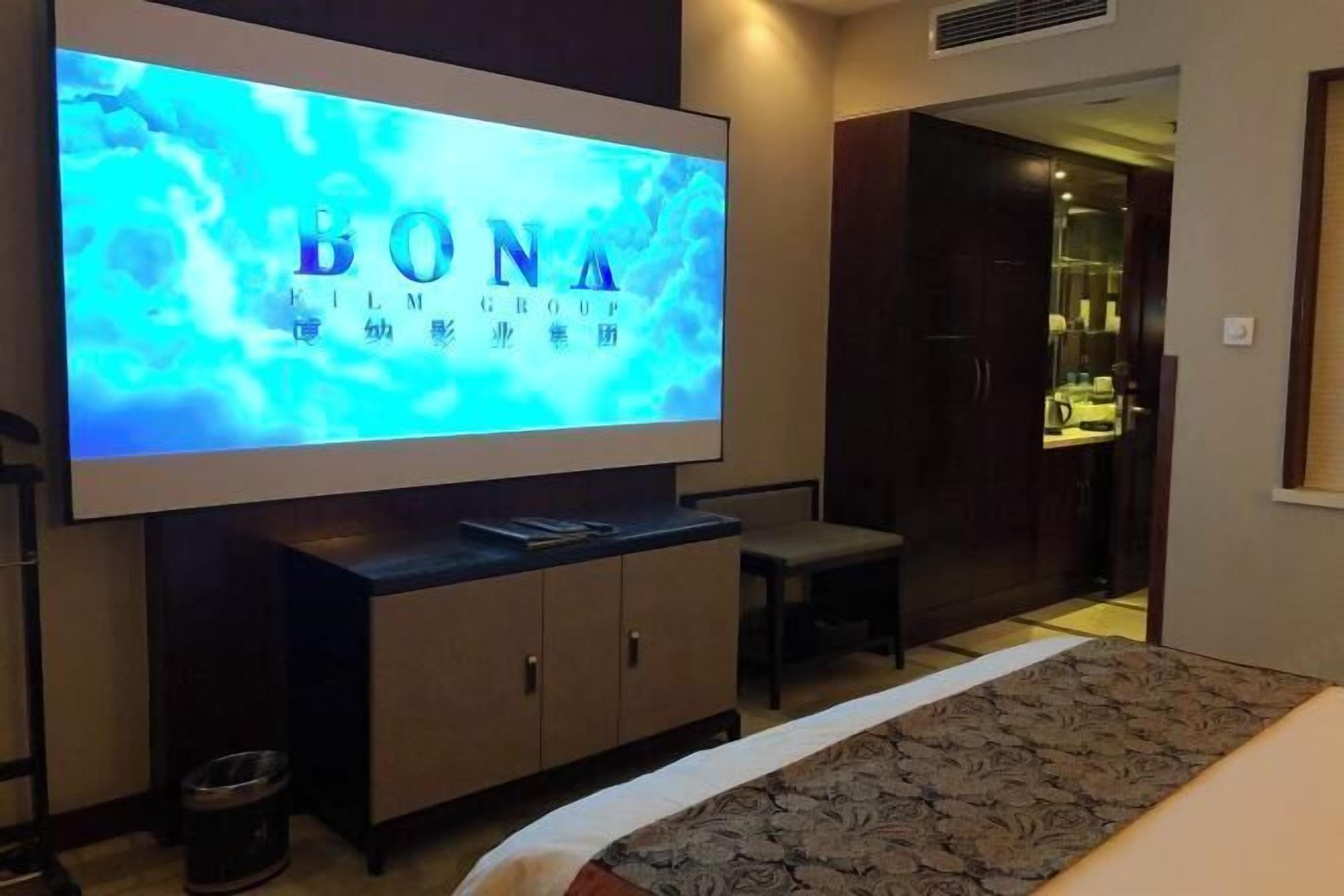 A hotel room with a large flat-screen TV displaying 'BONA FILM GROUP' on the wall. There is a bed with a patterned bedspread and a small bedside table with a lamp.