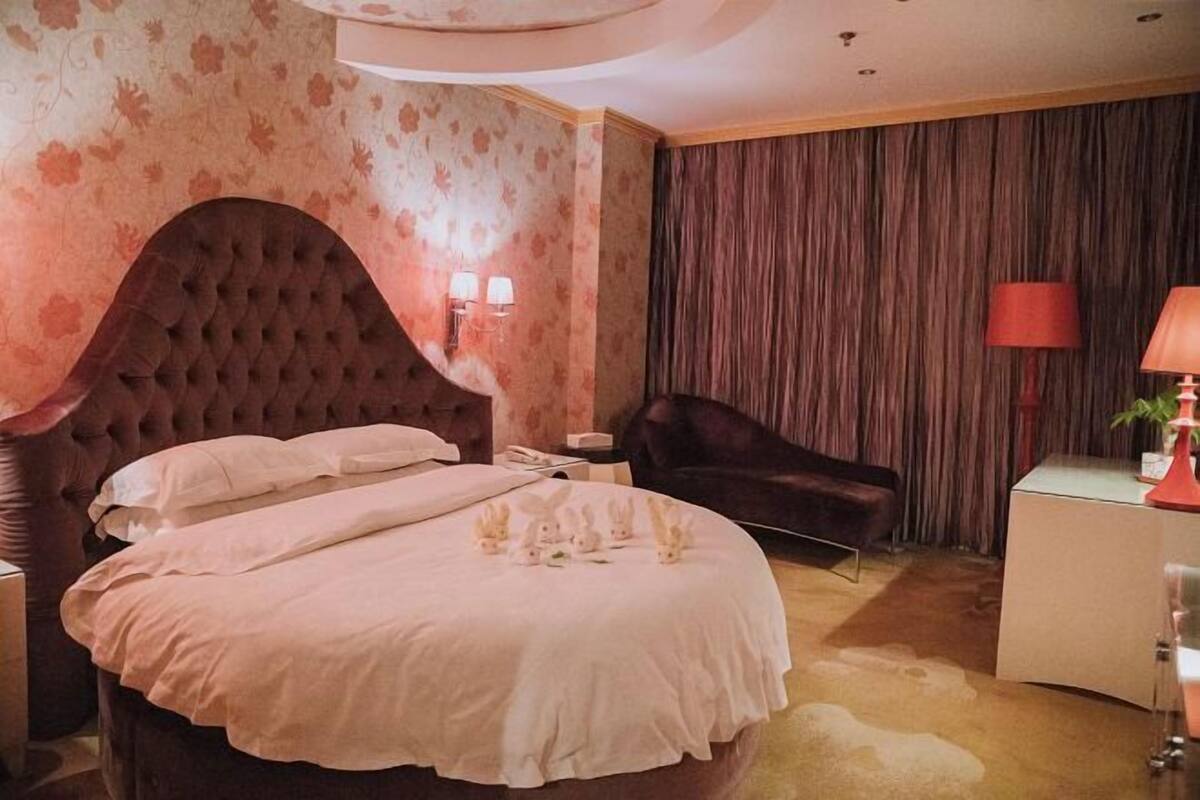 Een slaapkamer met een groot, gestoffeerd hoofdeinde, een bed met witte lakens, een donkere fauteuil, een nachtkastje met een lamp, en een raam met gordijnen.