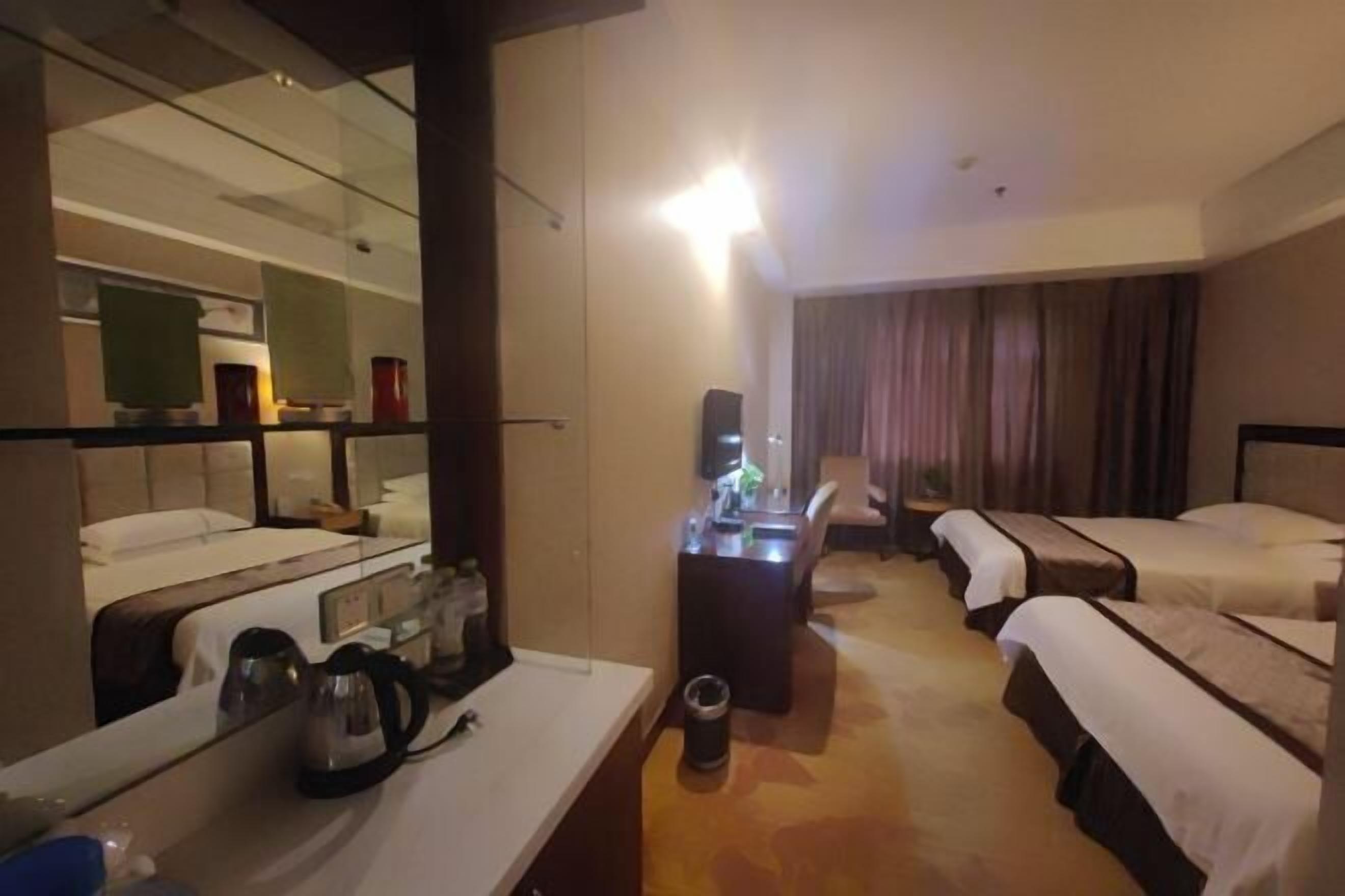 A hotel room with a large mirror, a desk with a kettle, two beds, a small table, and a chair.