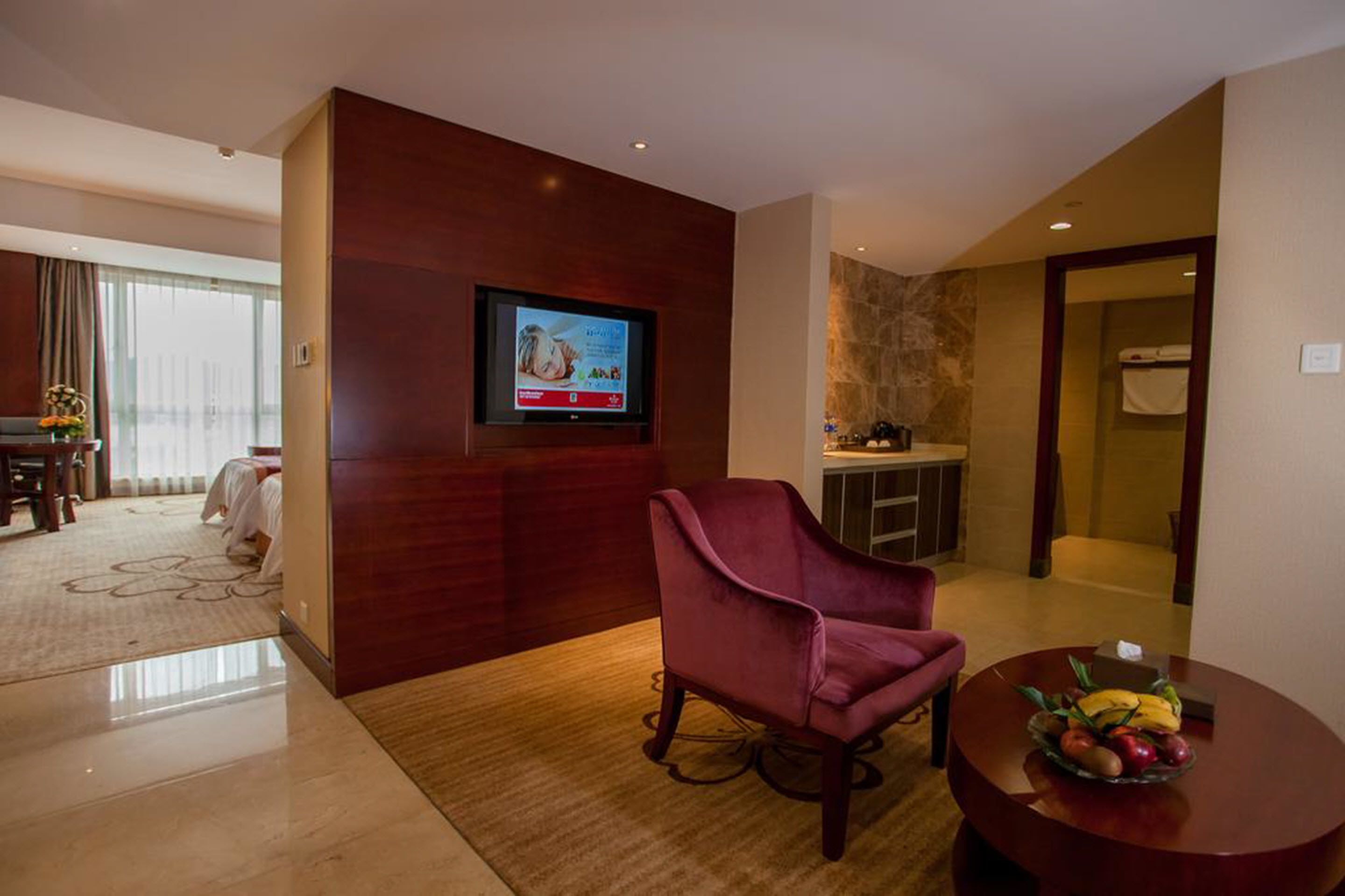 A hotel room with a flat-screen TV mounted on a wooden wall, a plush armchair, a round wooden table with a fruit bowl, and a glimpse of a bathroom with a towel rack.