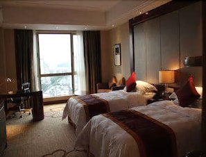A hotel room with two beds, a desk, a chair, and a large window with curtains. - Ramada Plaza Shaoguan City Centre (Shaoguan)