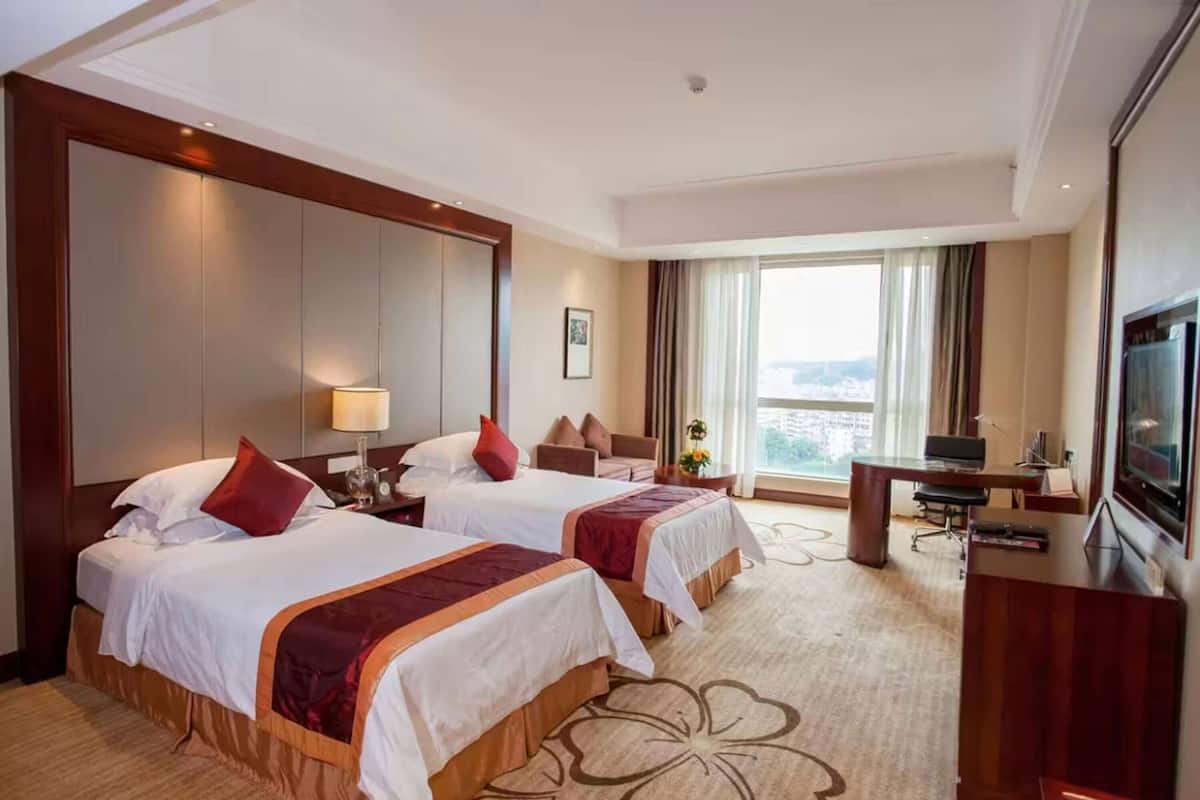Habitación de hotel con dos camas, un escritorio, un televisor y un ventanal con vista a la ciudad.