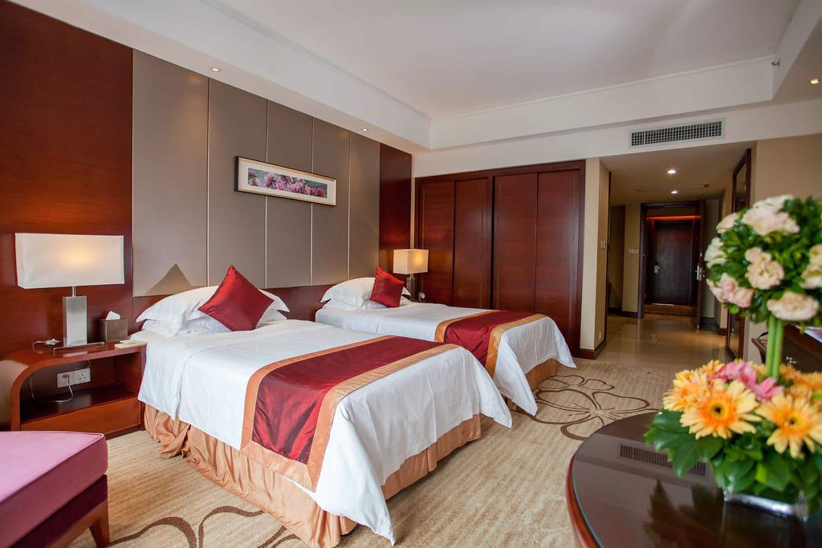 A hotel room with two beds, a desk, a chair, a bedside table, a lamp, a picture on the wall, and a vase with flowers.