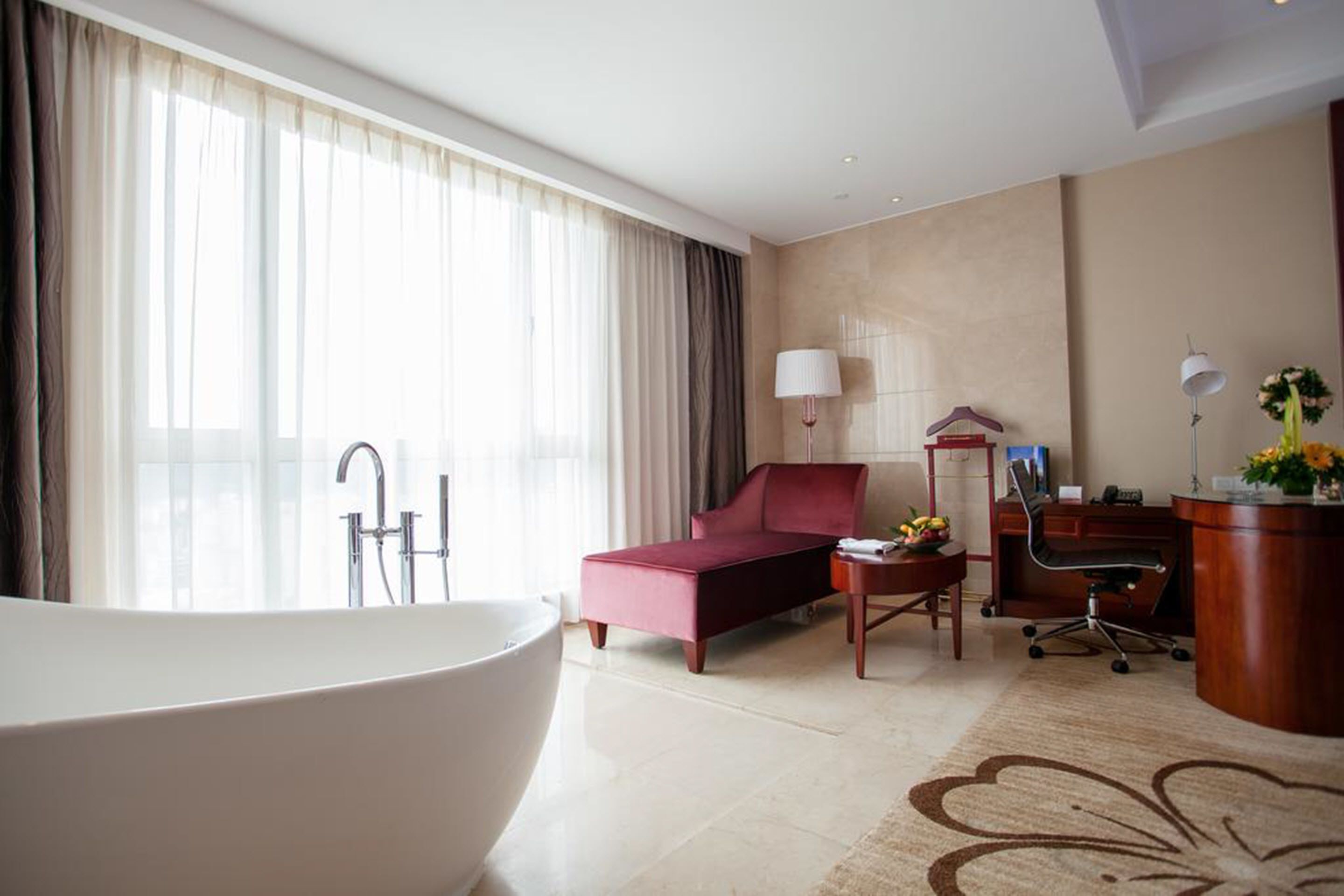 Una habitación de hotel moderna con una bañera grande, un sofá rojo, un escritorio con una silla y un ventanal con cortinas.