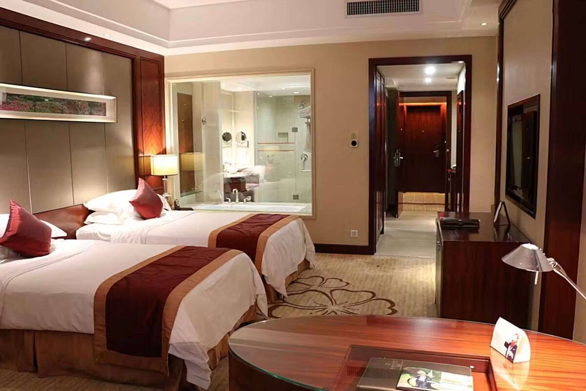 Habitación de hotel con dos camas, un escritorio, un televisor y un baño visible a través de una puerta de vidrio.