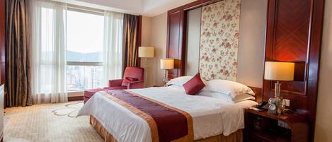 A hotel room with a large bed, a desk, a chair, and a view of the city through the window.