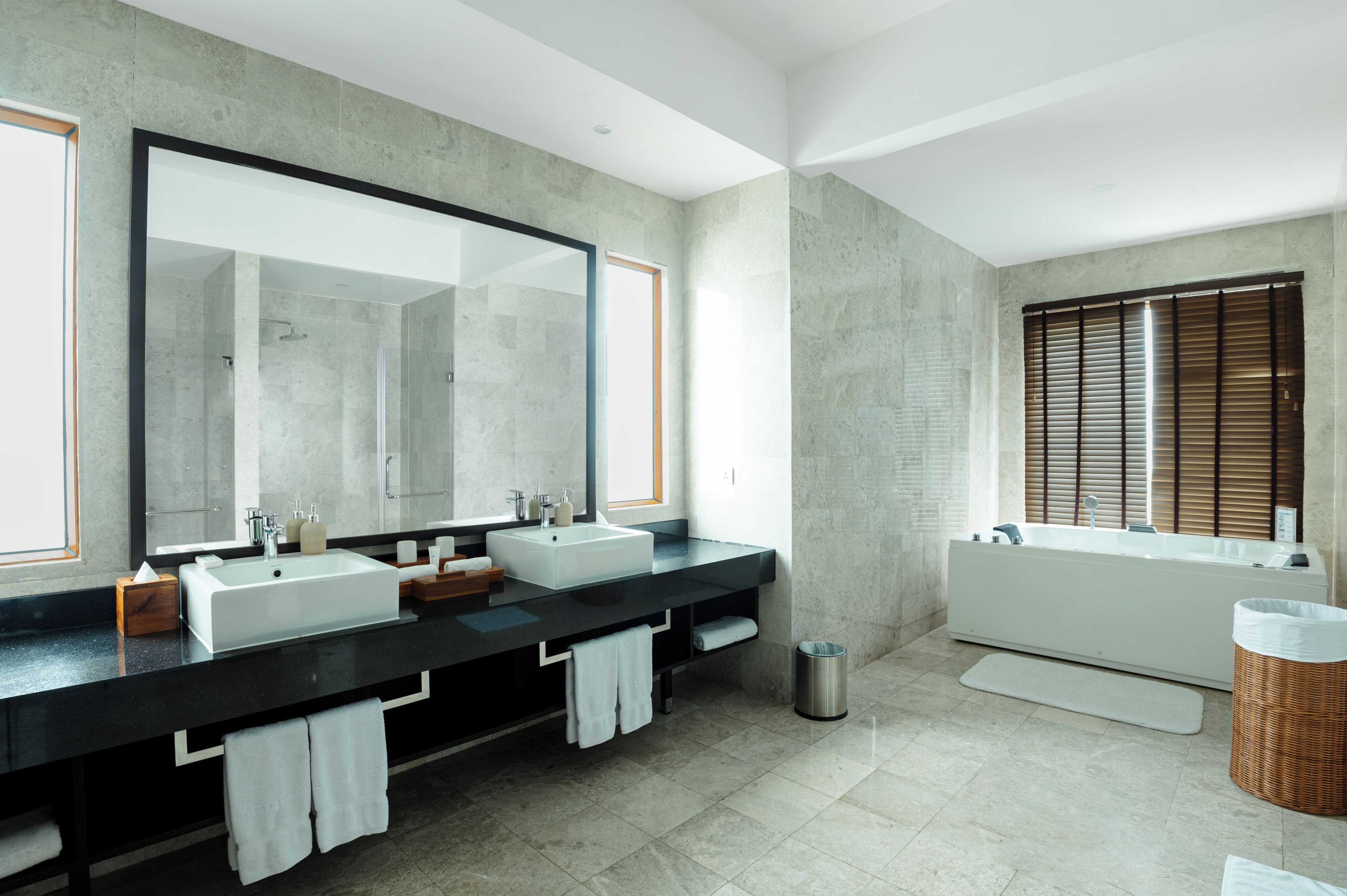 A modern bathroom with two sinks, a large mirror, a bathtub, and a window with blinds.