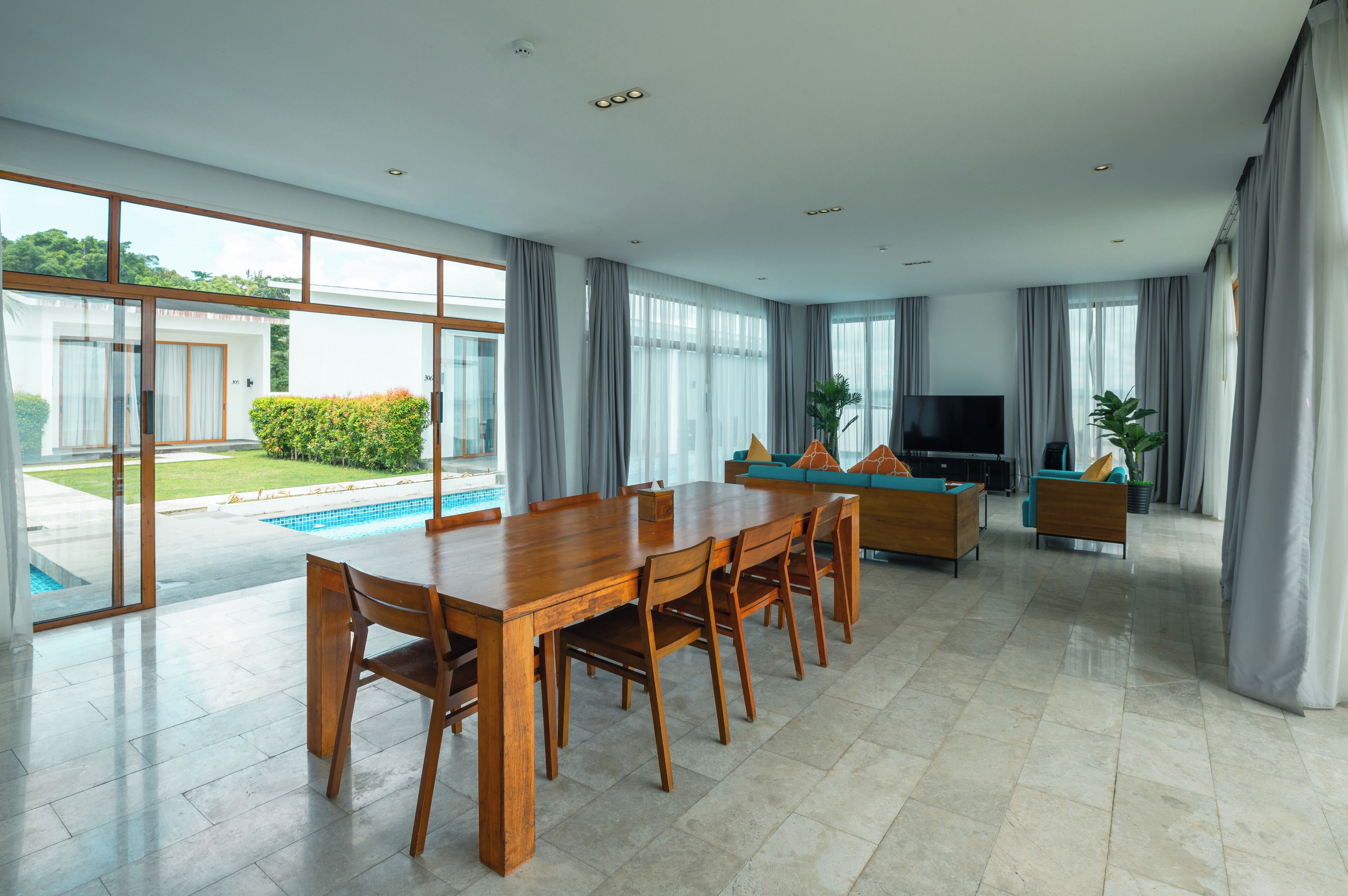 A spacious living area with a large wooden dining table, comfortable seating, a flat-screen TV, and a view of a swimming pool and greenery outside.