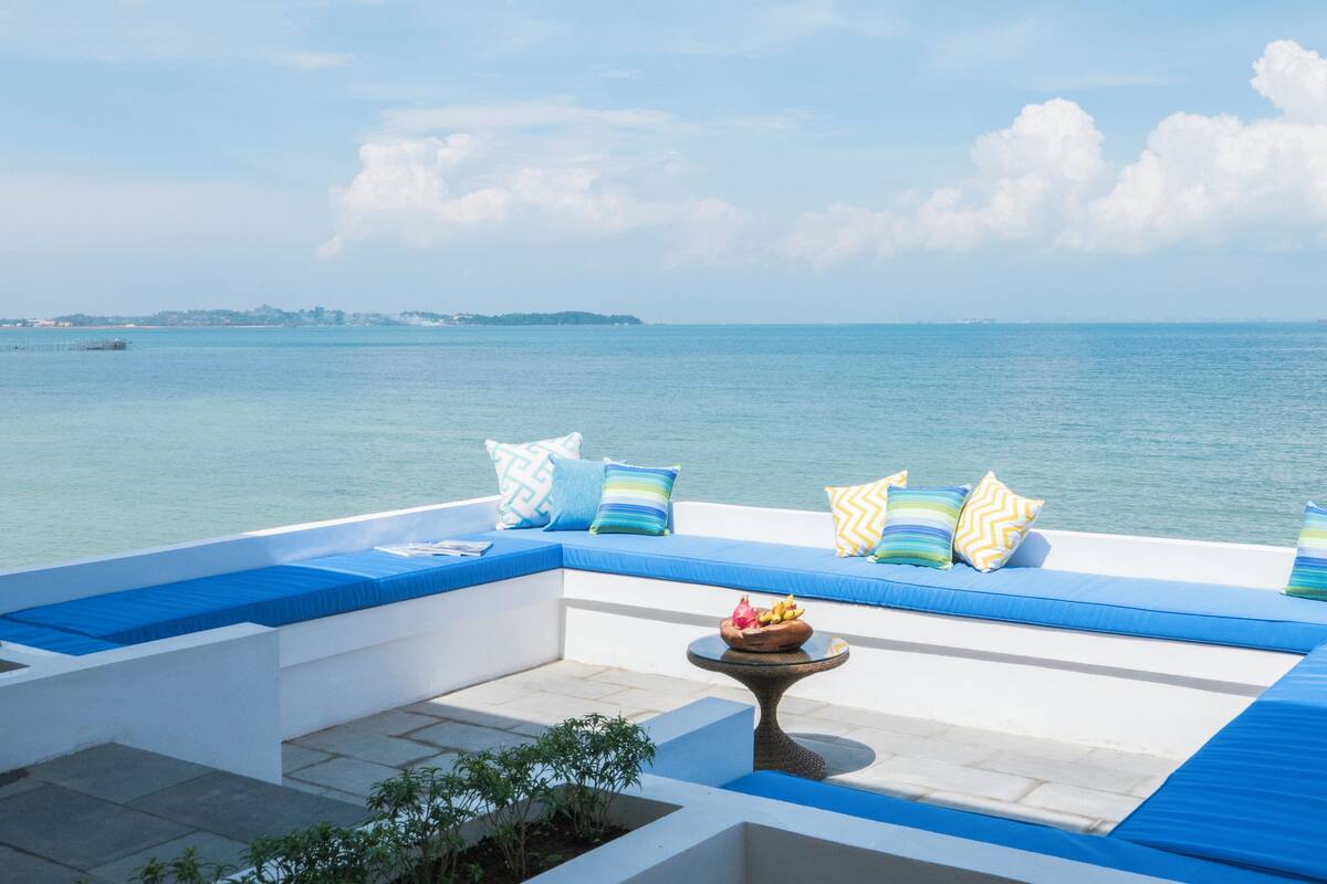 Ein moderner Außenbereich mit Sitzgelegenheiten, blaue Kissen und gemusterte Zierkissen, mit Blick auf ein ruhiges Meer.