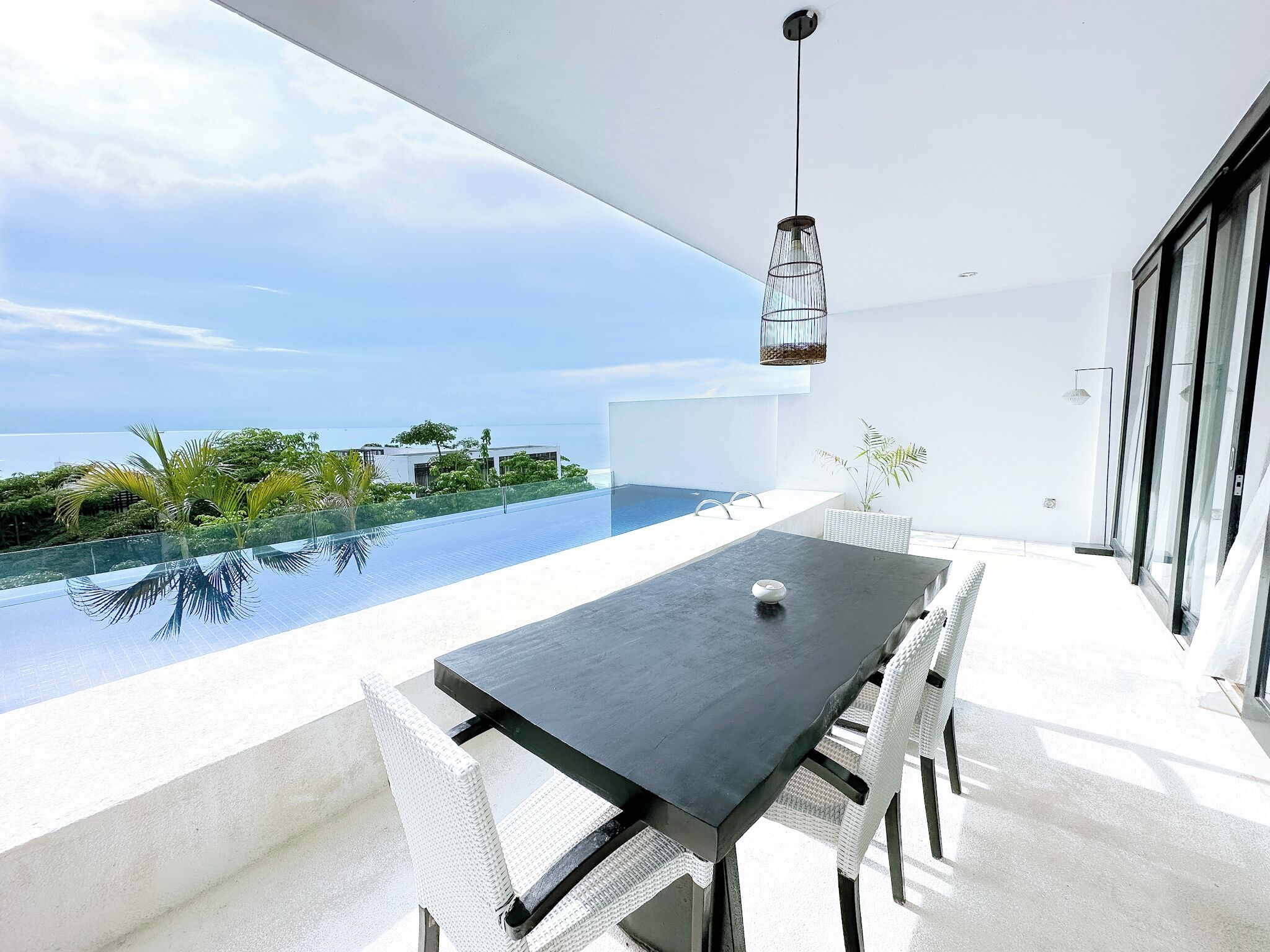 A modern outdoor dining area with a pool, white wicker furniture, and a hanging light fixture.
