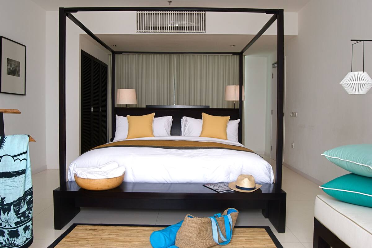 Ein Himmelbett mit Holzgestell, weißer Bettwäsche und gelben Kissen. Auf dem Bett liegt ein gewebter Korb, ein Hut und auf dem Boden eine blaue Tasche.