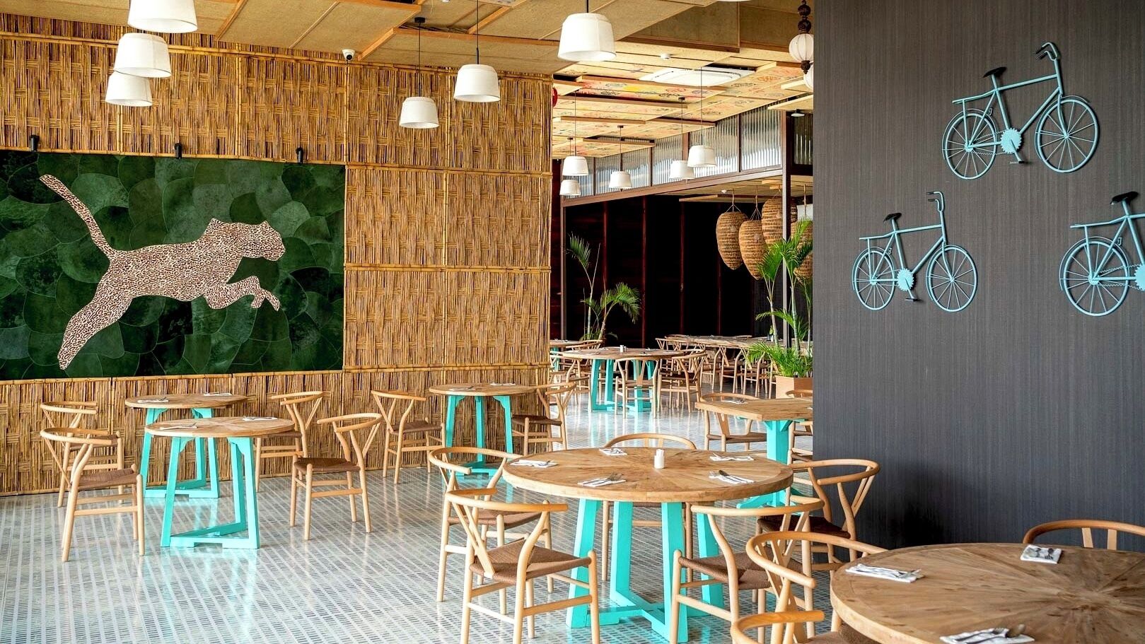 L’intérieur d’un restaurant est décoré de tables rondes, de chaises, de luminaires suspendus, d’une fresque représentant un léopard et d’éléments décoratifs sur le thème du vélo.