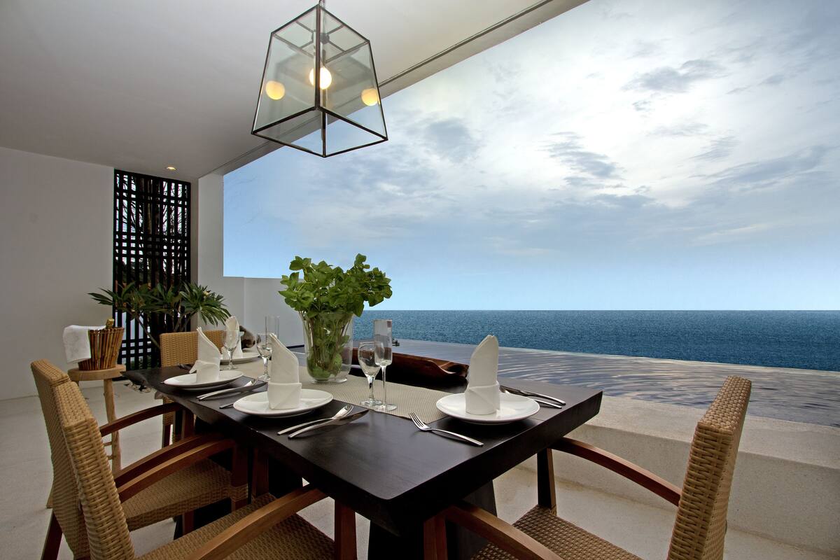 Ein Esstisch mit weißem Geschirr, Besteck und Gläsern, mit Blick auf das Meer.