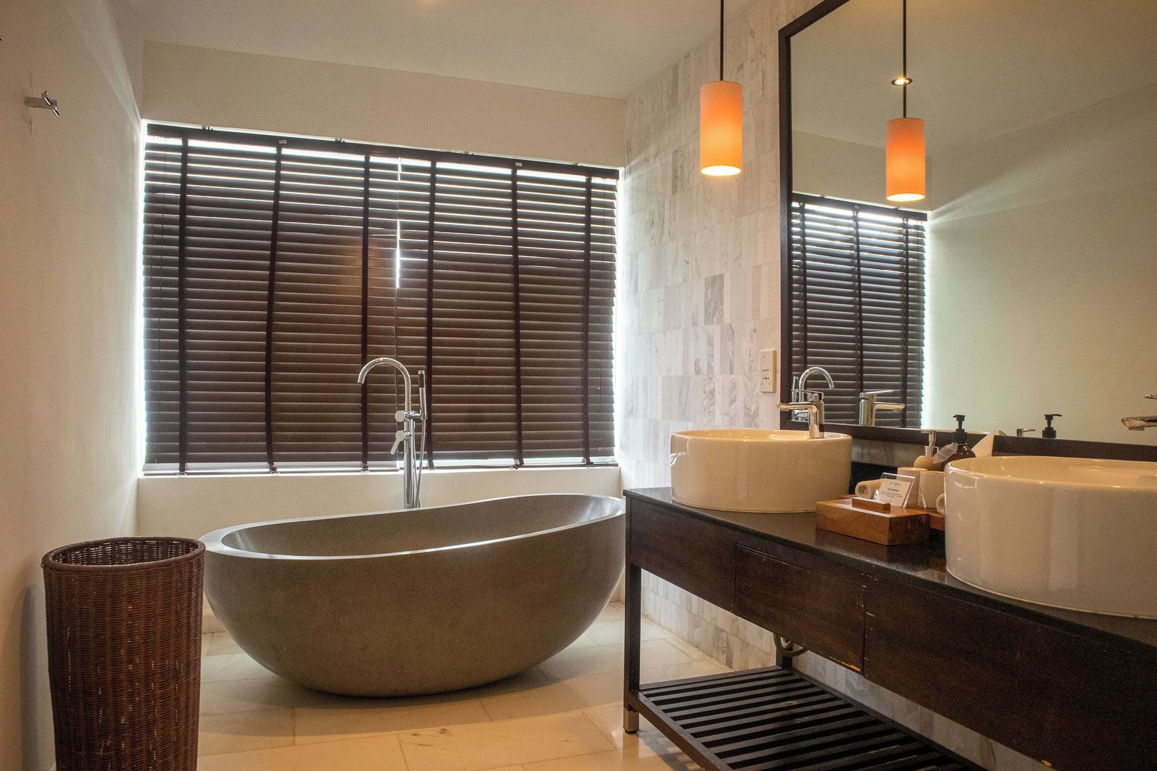 Een moderne badkamer met een vrijstaand ligbad, twee wastafels, een grote spiegel en een raam met zonwering.