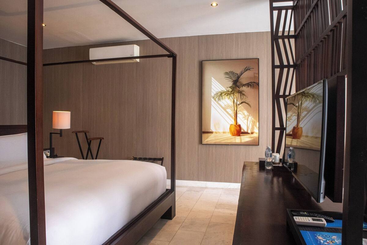 Ein modernes Hotelzimmer mit Himmelbett, Schreibtisch inklusive Computer, großem gerahmten Bild und Fernseher auf Holzfuß.