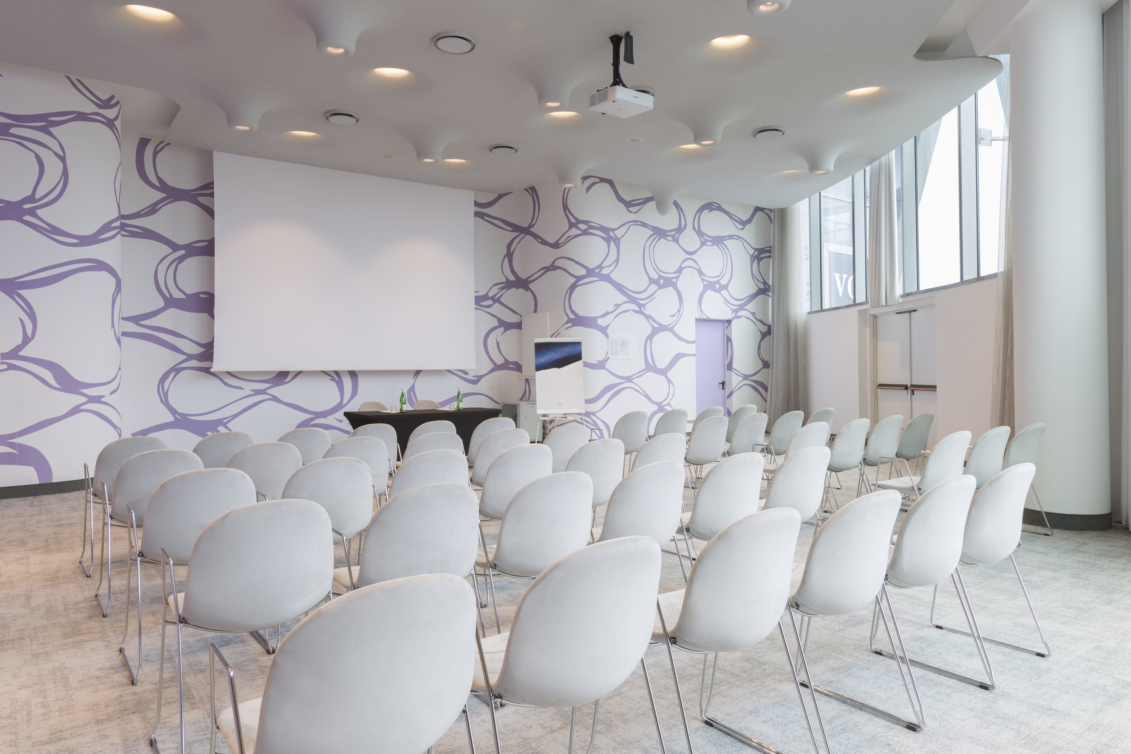 A conference room with rows of white chairs, a large white screen, and a projector.