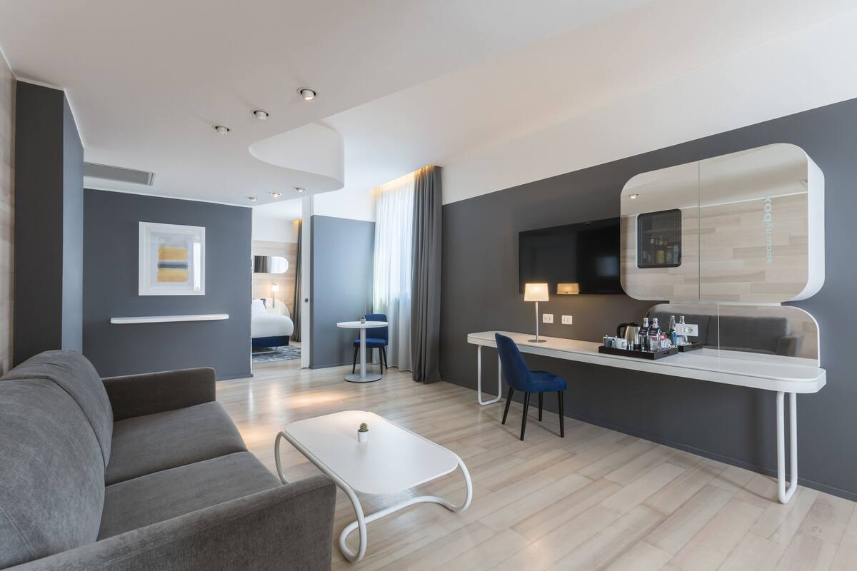 A modern hotel room with a grey sofa, a white coffee table, a desk with a chair, and a large mirror.