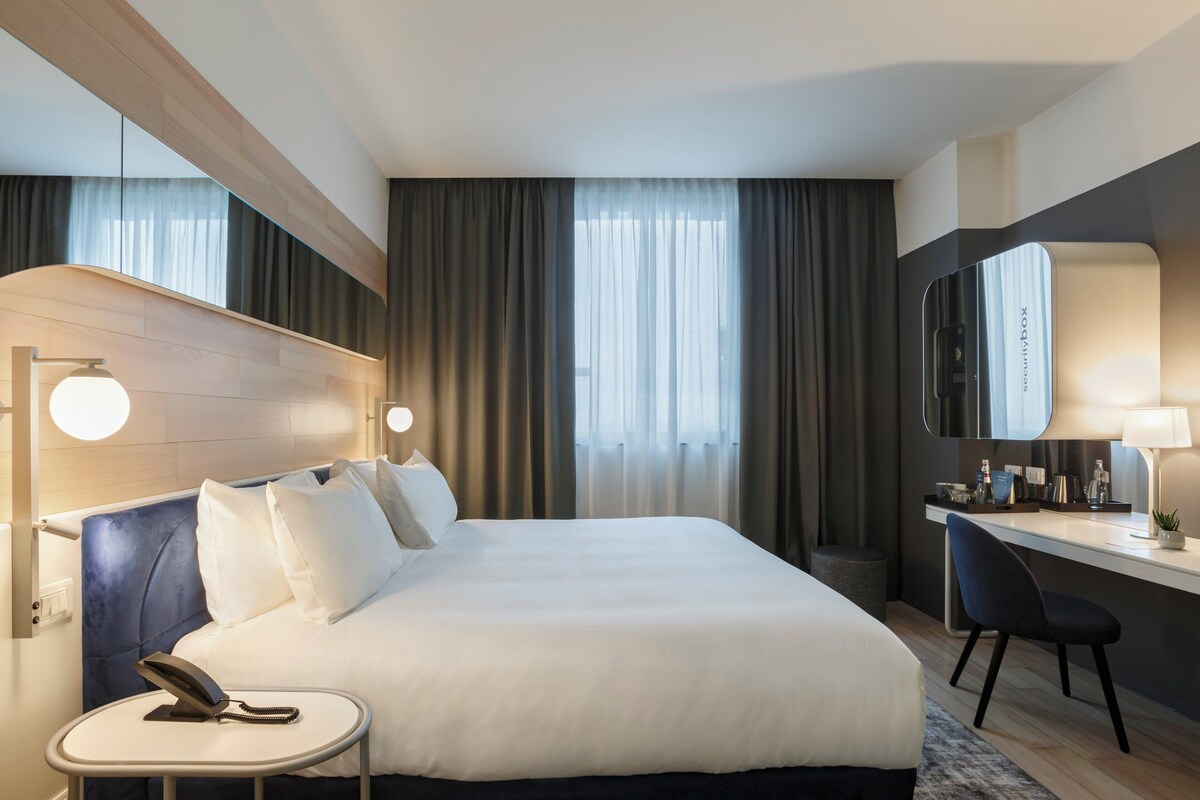 A hotel room with a large bed, bedside table with a phone, a desk with a chair, a mirror, and curtains.