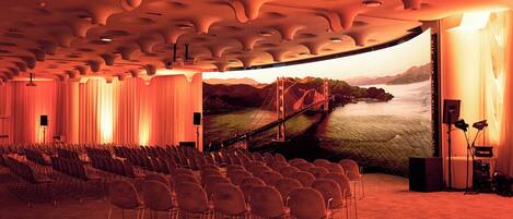 A conference room with rows of chairs, a large screen displaying a scenic view, and a stage with audio equipment.