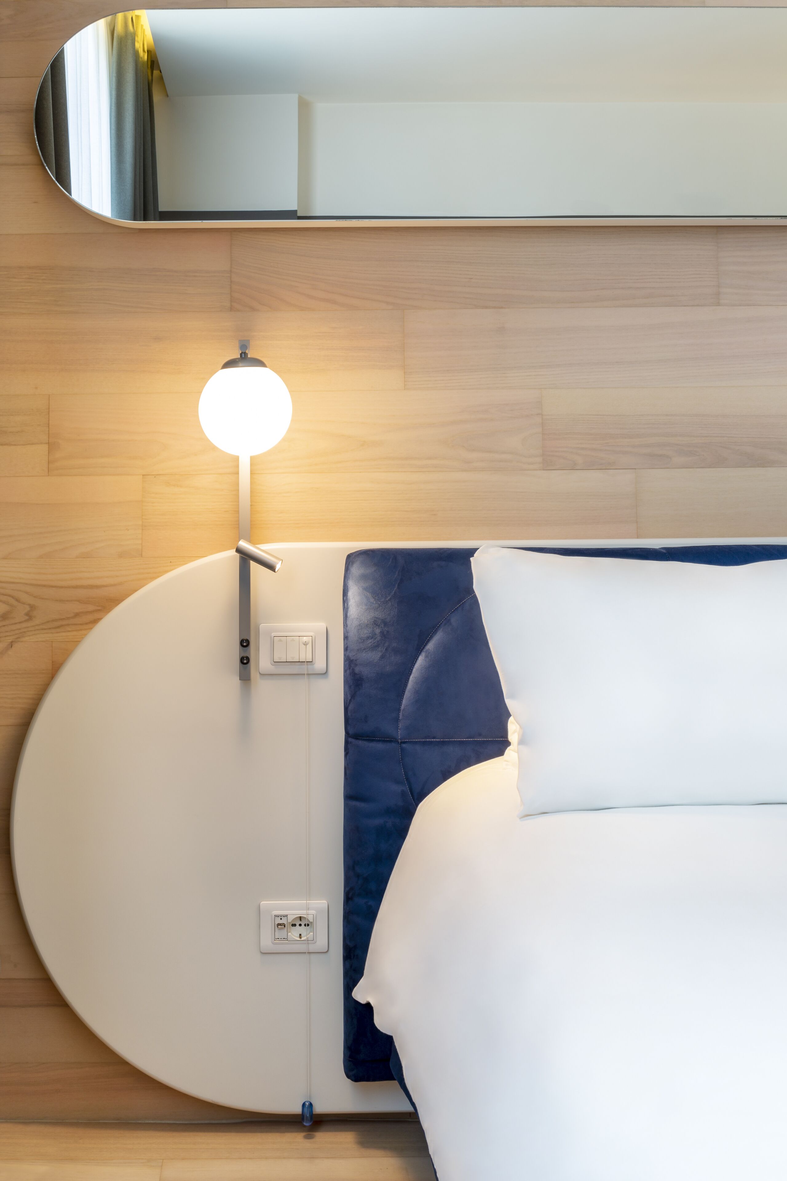 A modern hotel room with a round bed, a bedside table with a lamp, and a large mirror above the bed.