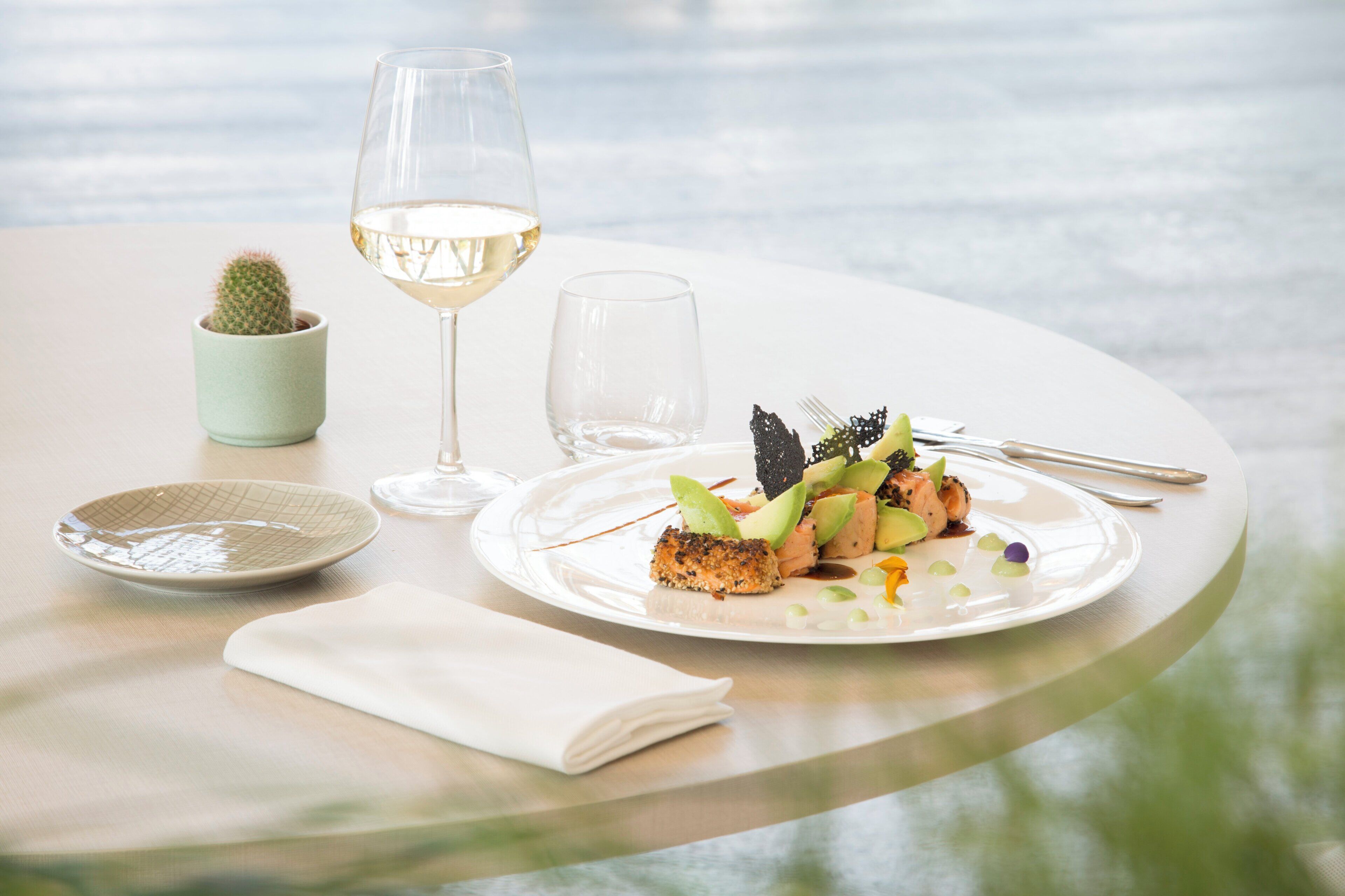 A table with a plate of food, a glass of white wine, a small cactus, and a folded napkin.