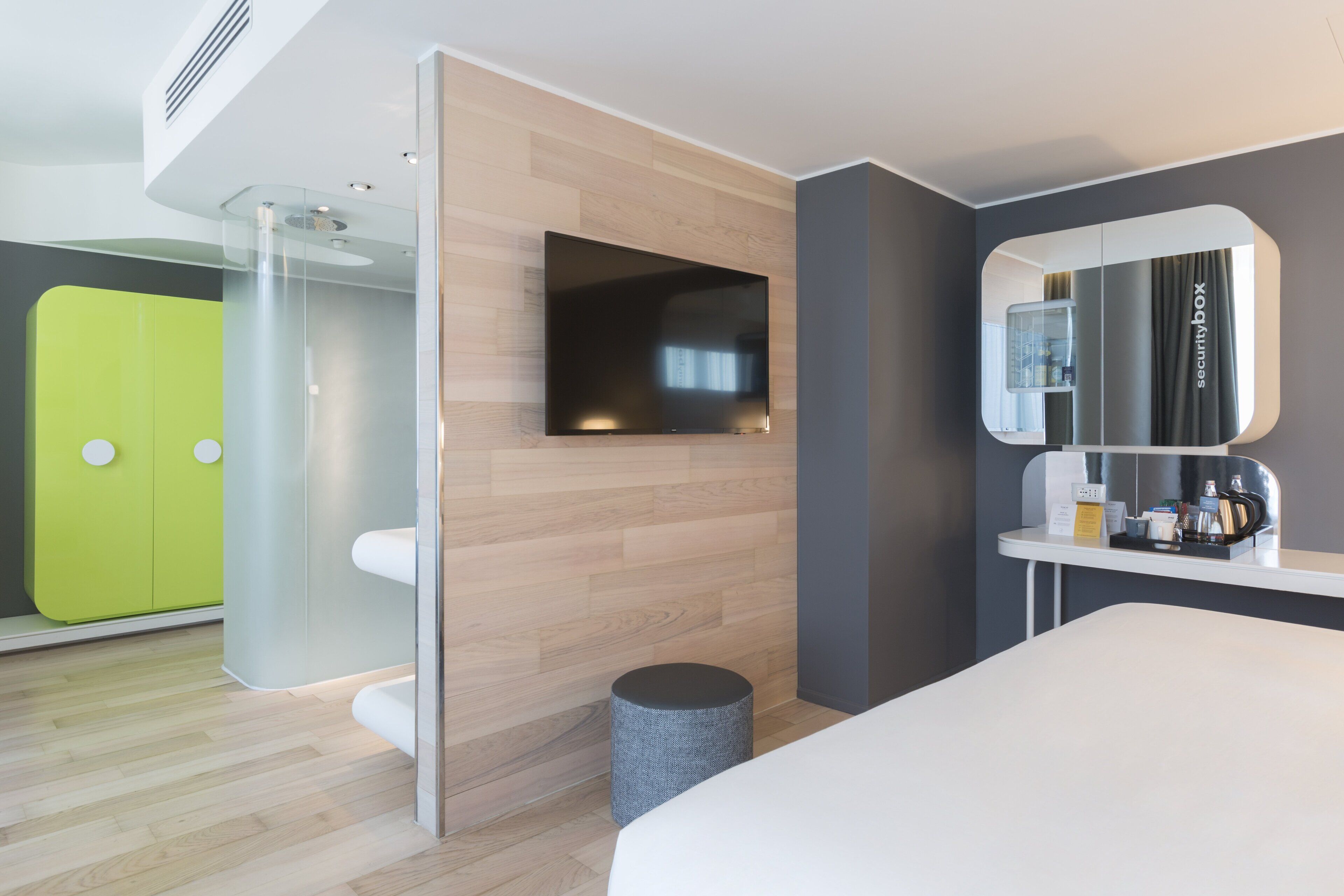 Ein modernes Hotelzimmer mit einem großen Bett, einem Flachbildfernseher an einer Holzwand, einem kleinen runden Hocker und einem Spiegel mit Hinweis auf einen Safe.