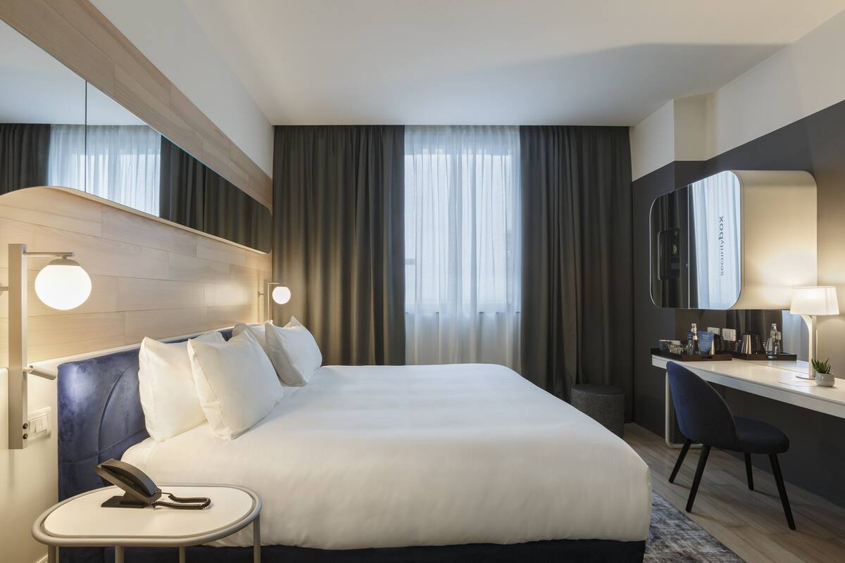 A hotel room with a large bed, bedside table with a phone, a desk with a chair, a mirror, and curtains.