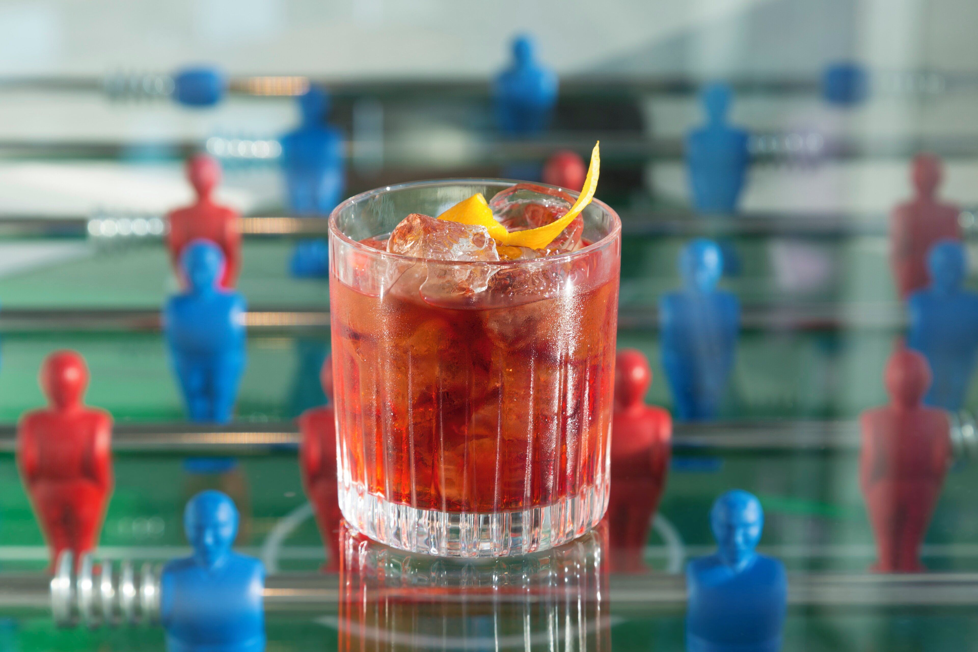 A glass with a red beverage, ice, and a lemon slice on a reflective surface.